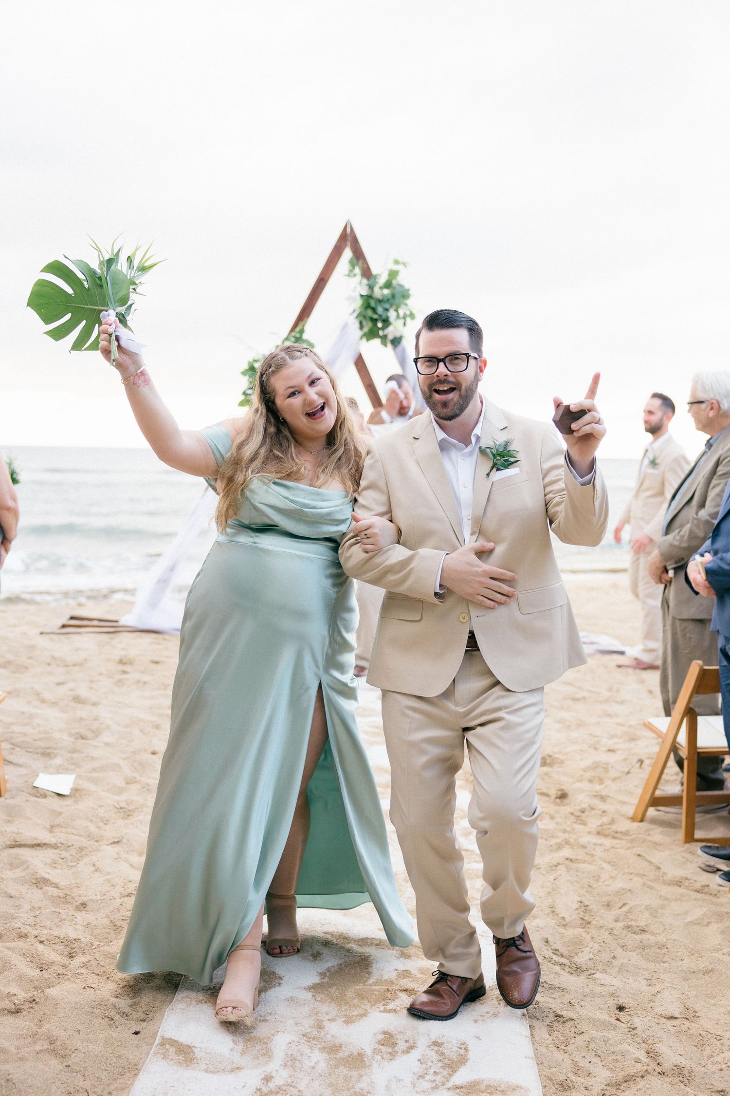 Wedding party celebrates joyfully on beach with tropical leaf props and mint green attire.