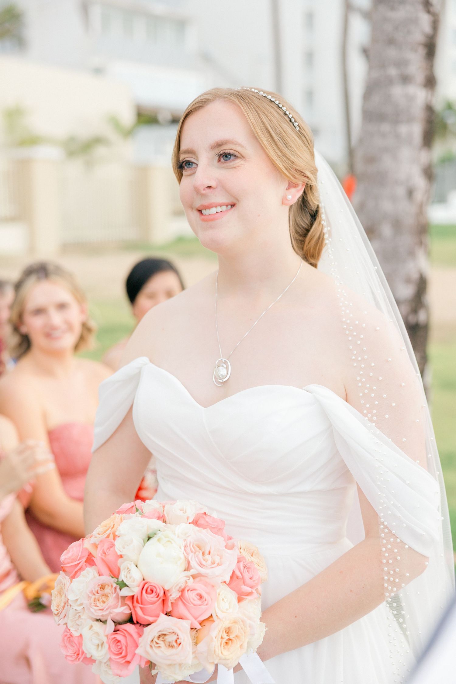 Bride holds pink and peach rose bouquet during outdoor wedding ceremony.