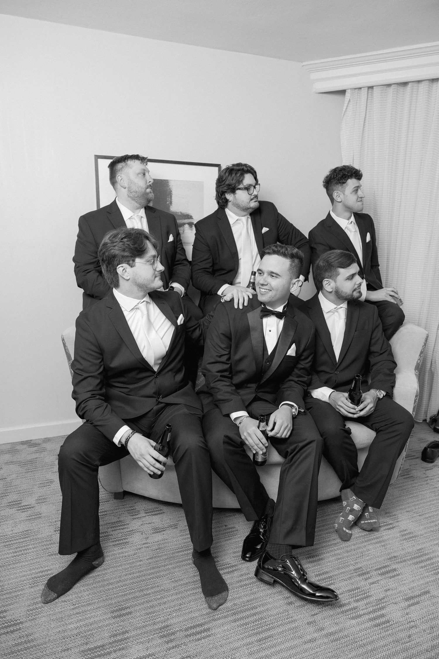 Group of groomsmen in black tuxedos sitting together on a couch at formal event.