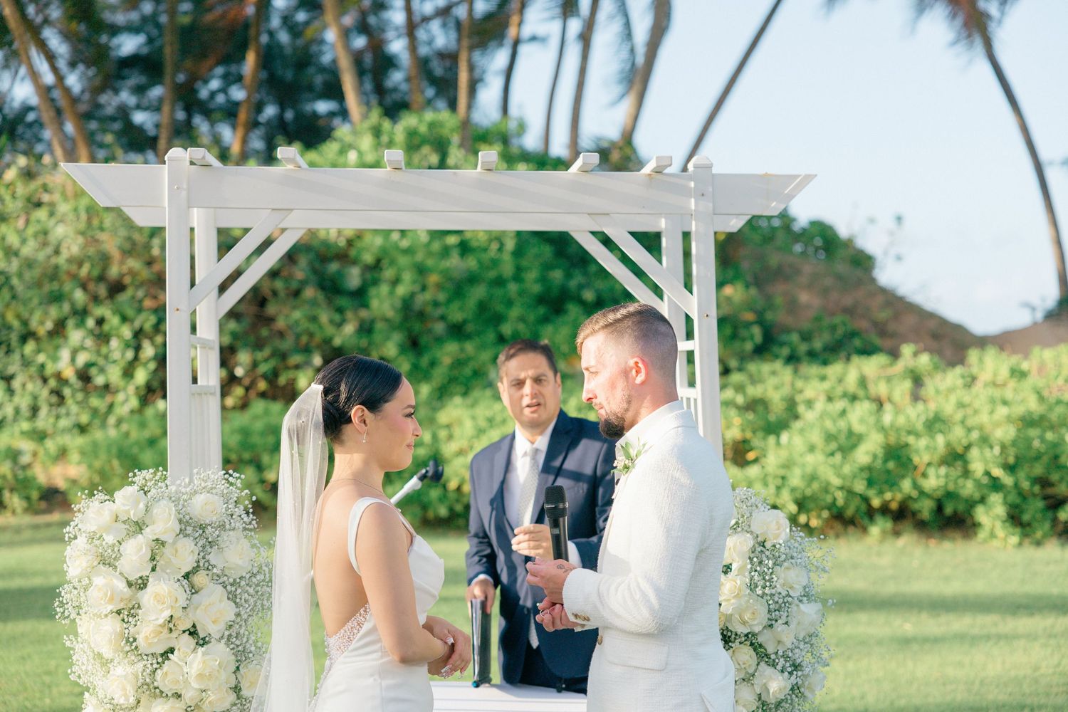Outdoor wedding ceremony setup with white floral arrangements and tropical palm trees.