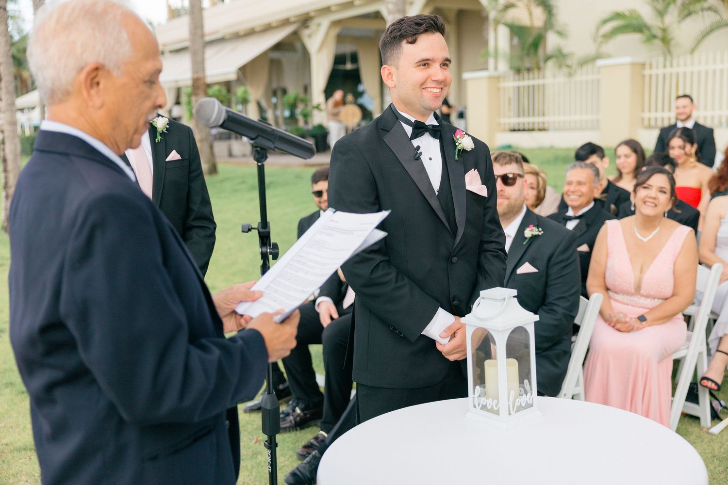 Wedding officiant leads outdoor ceremony as attendees in formal attire look on.