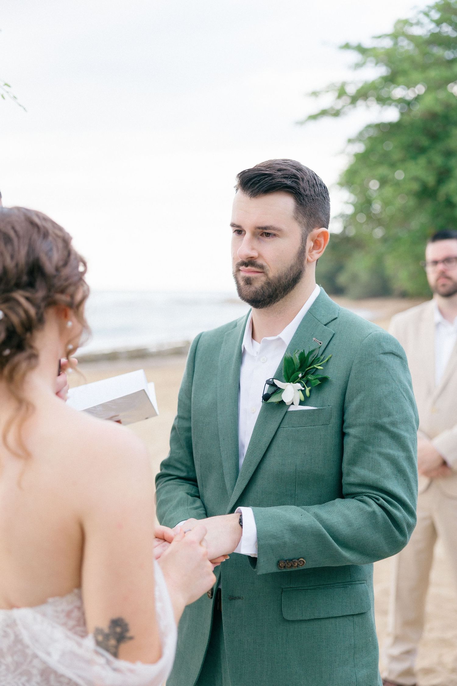 Intimate beach wedding ceremony with groom in green suit and bride in white lace dress.