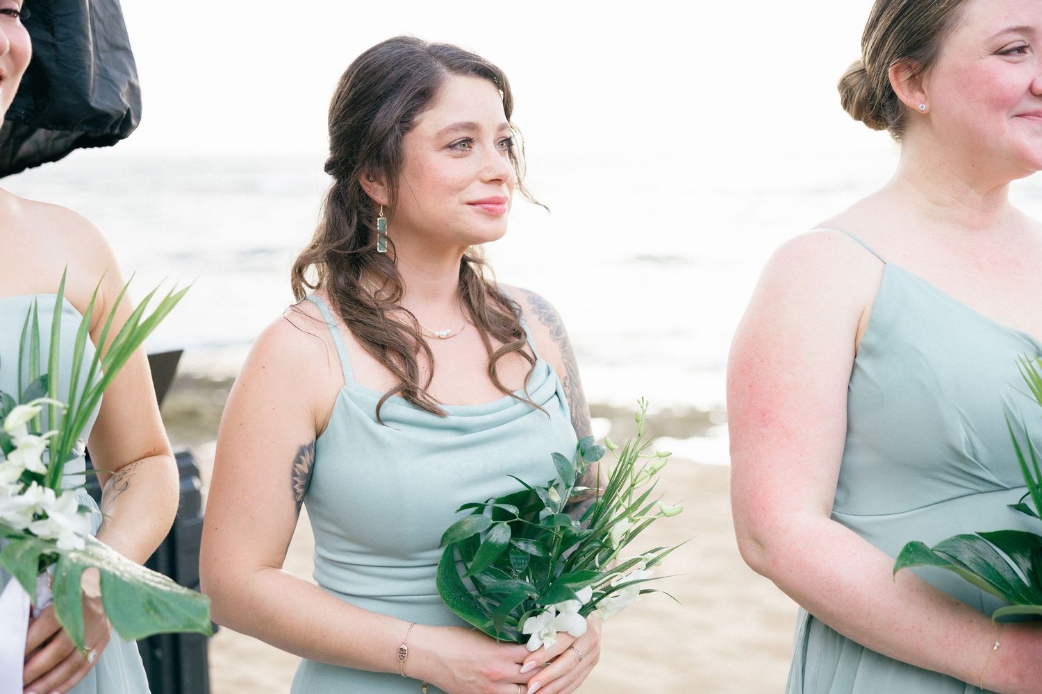 Bridesmaids in sage green dresses holding bouquets at beach wedding.
