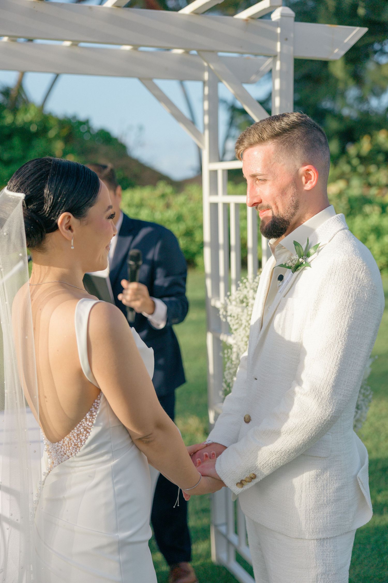 Wedding ceremony participants in white attire under white pergola structure.