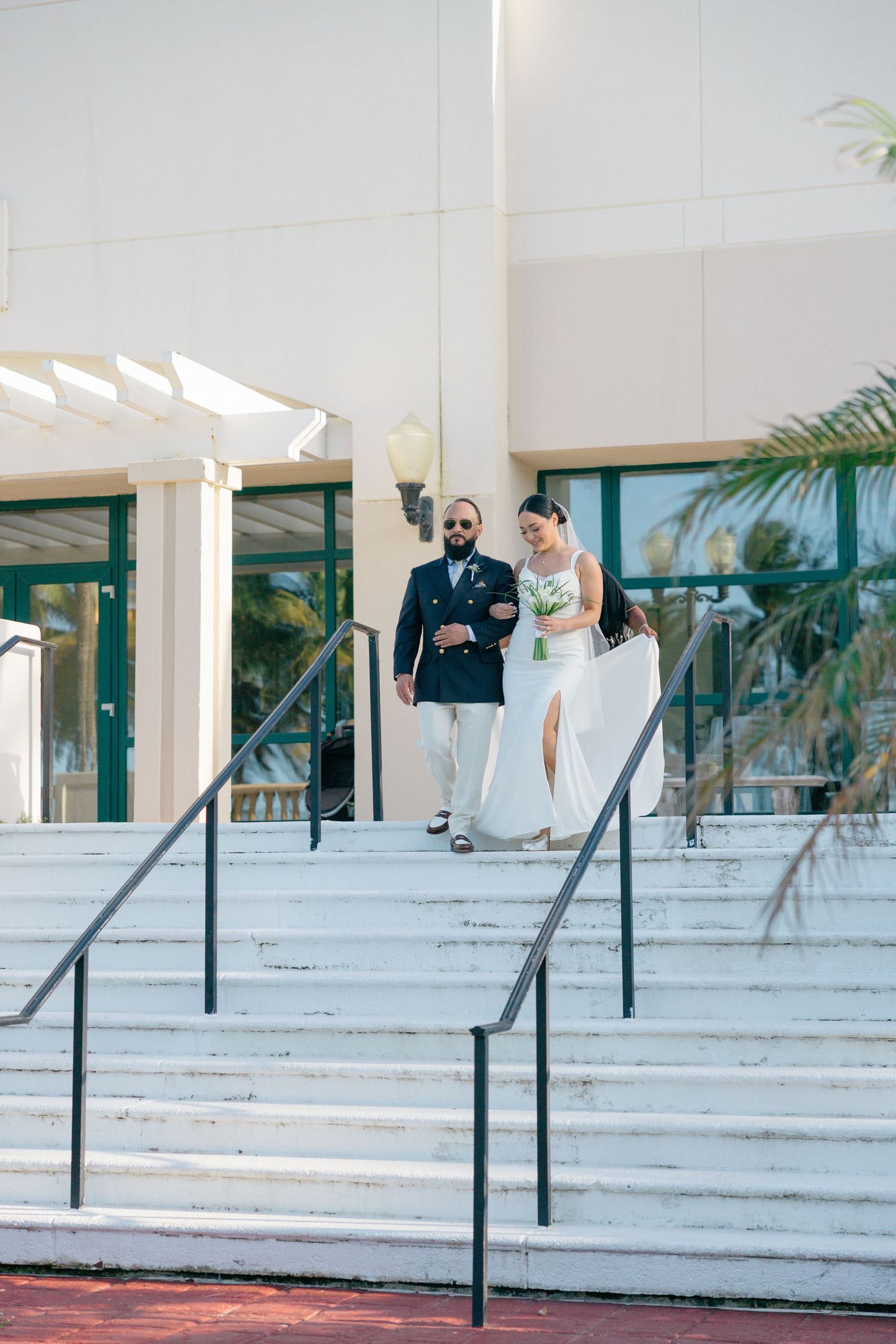 Modern white building entrance with steps and metal railings surrounded by palm trees.