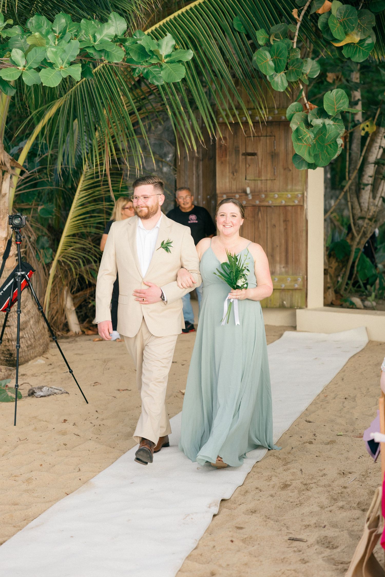 Wedding party members in light colored attire walk down a white aisle runner on a sandy beach.