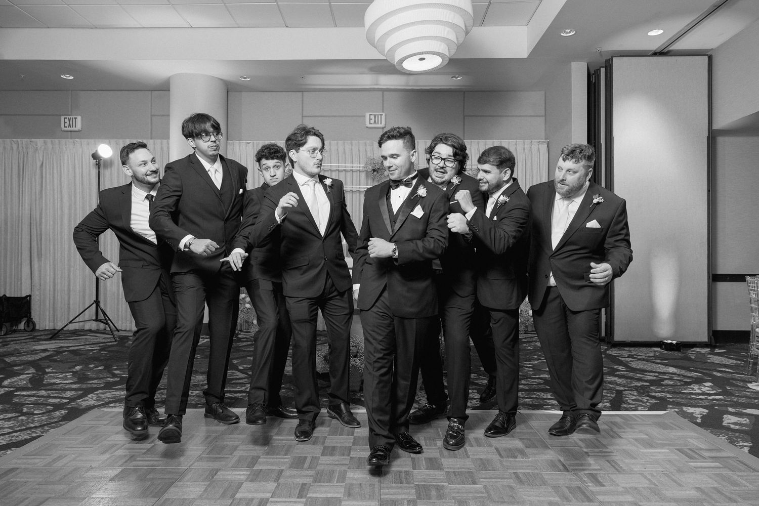 Wedding party in black suits poses energetically in a hotel ballroom.