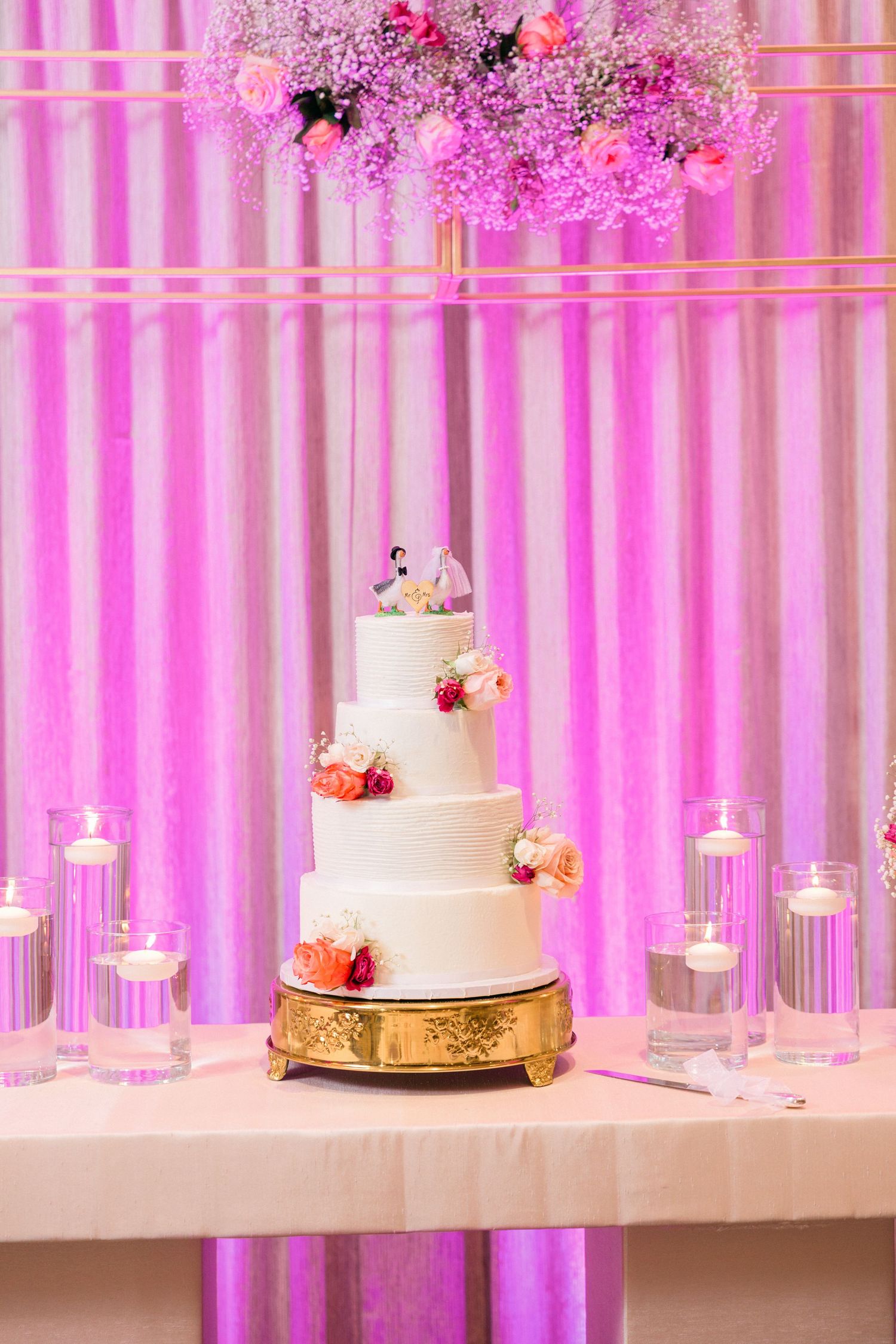 Wedding cake display table with pink uplighting and hanging floral arrangements.