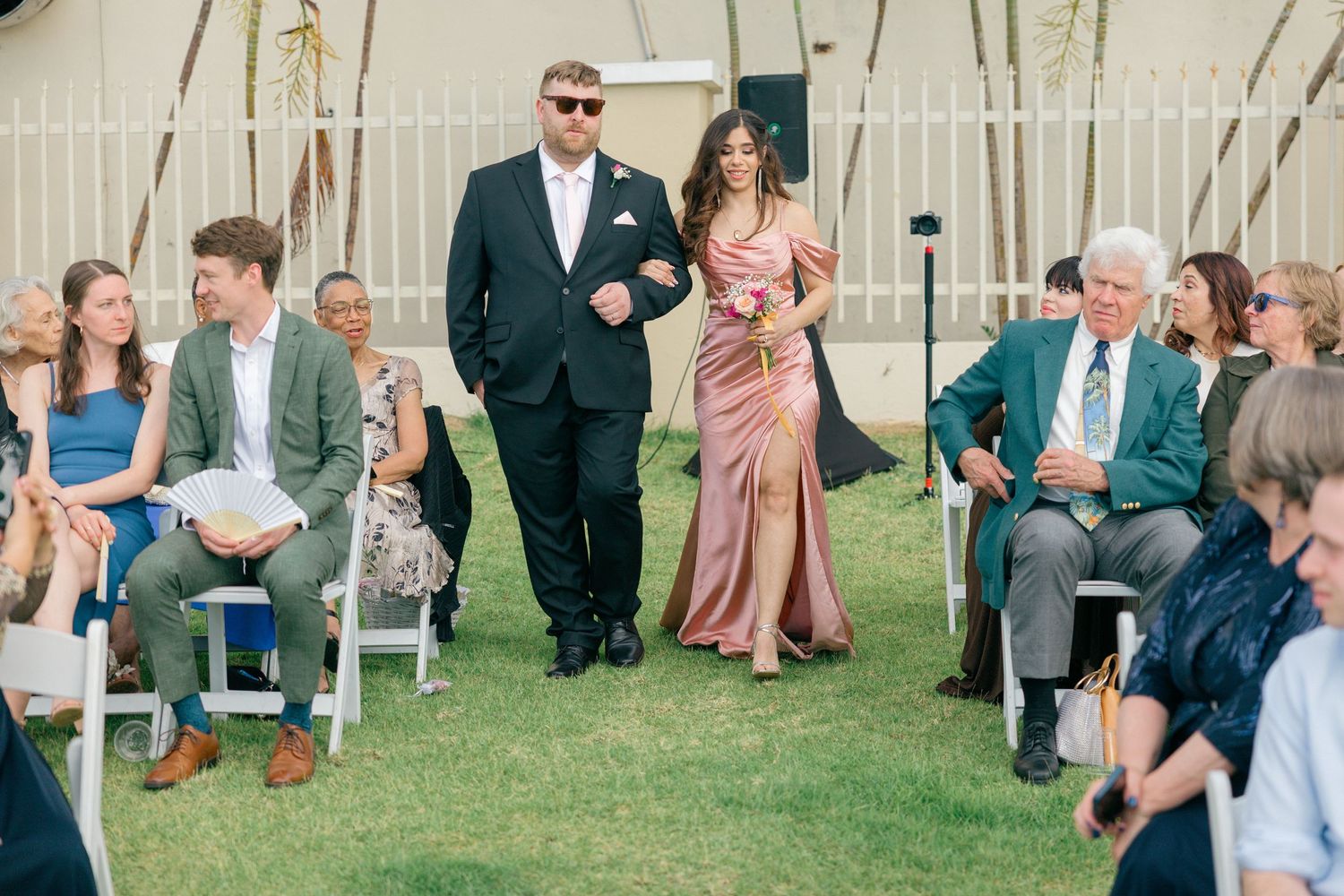 Outdoor wedding ceremony with guests seated on white chairs surrounded by tropical palm trees.