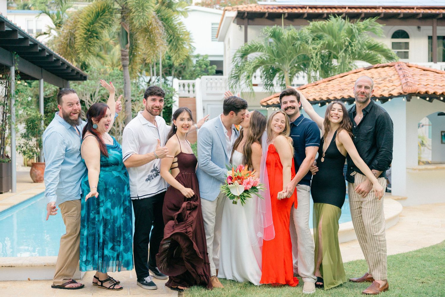 Wedding party members gather outdoors near a swimming pool wearing mix-and-match dresses in tropical colors.