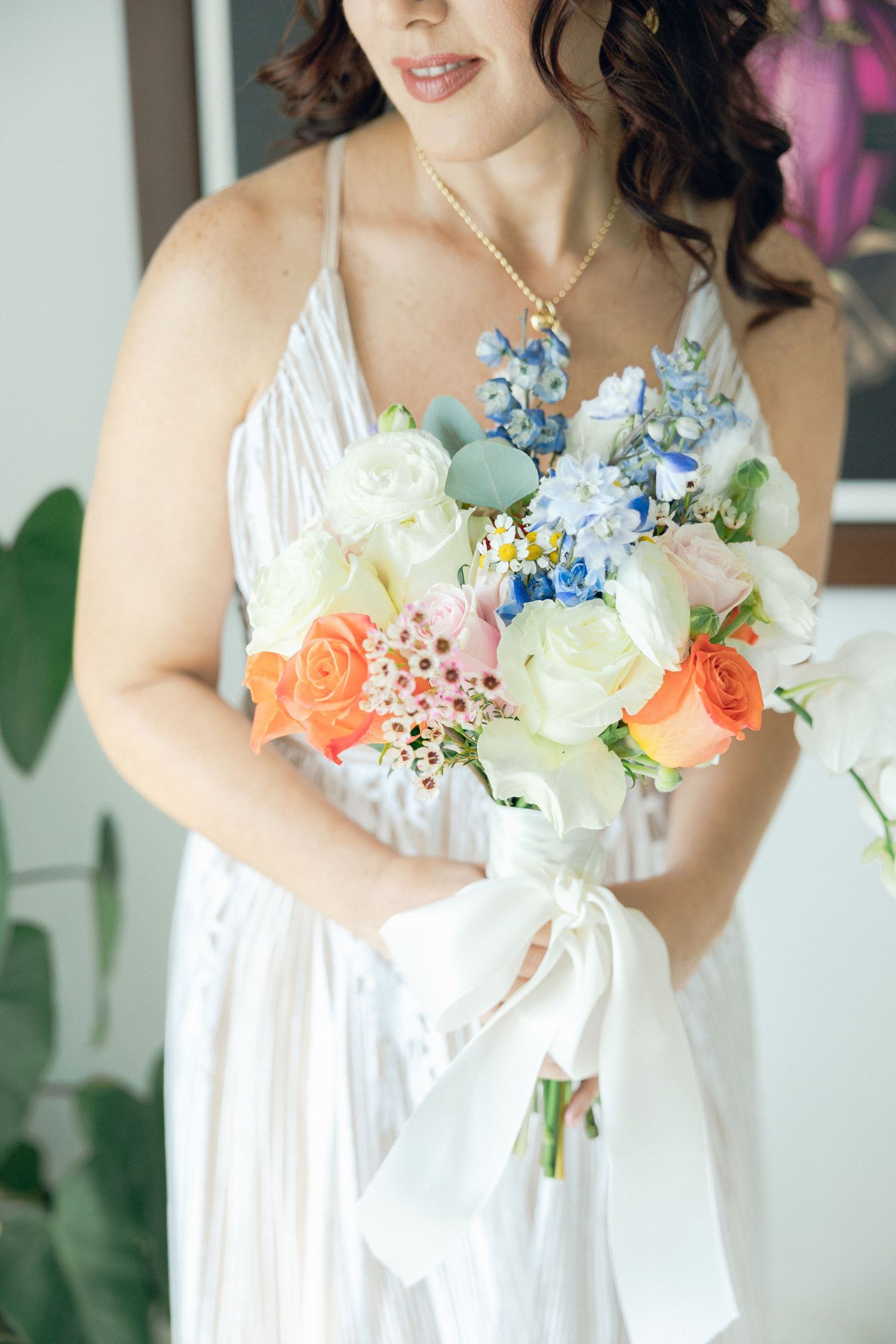 Close up of a bridal bouquet with white roses, coral flowers, and blue accents against a white dress.