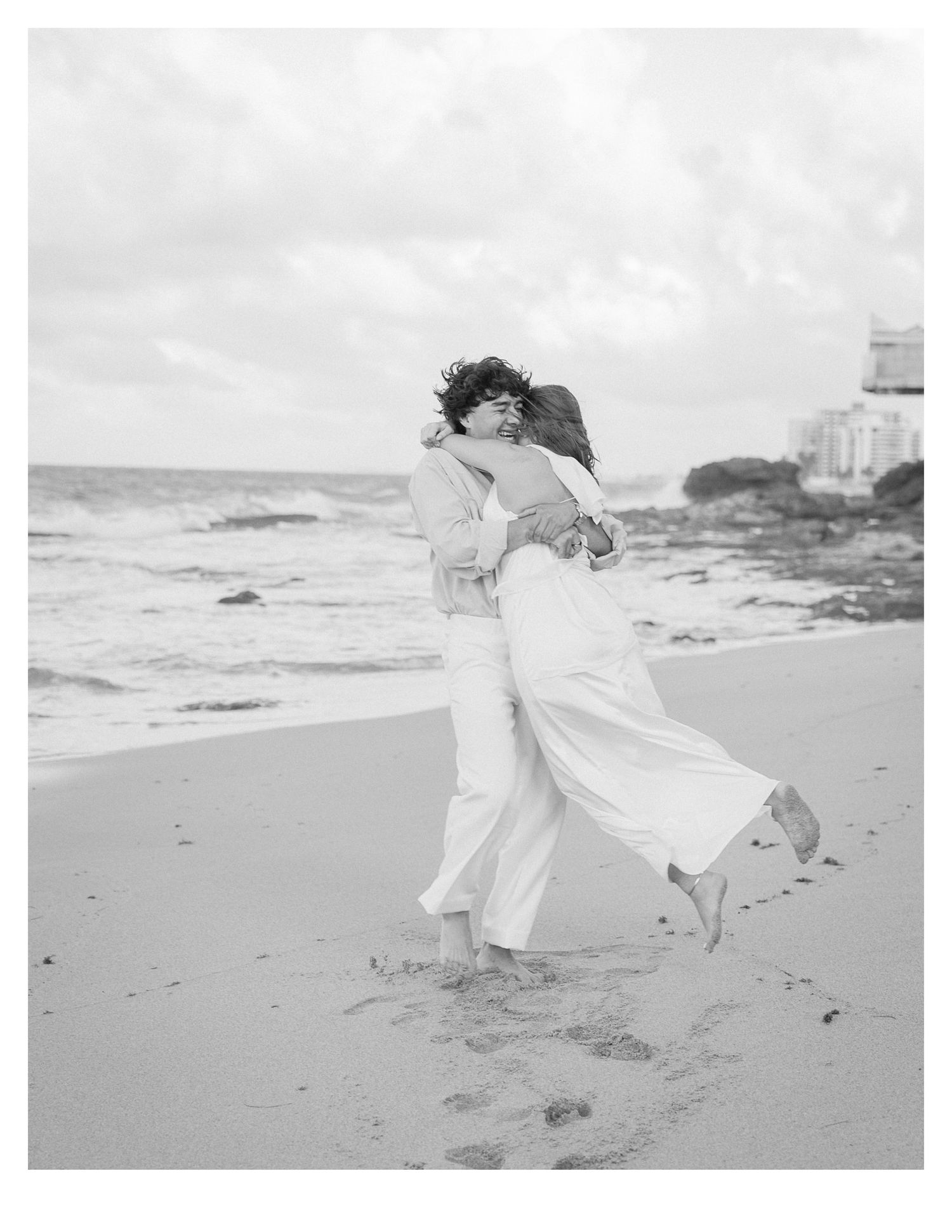 Black and white photo of a couple dancing on a beach with waves crashing behind them.