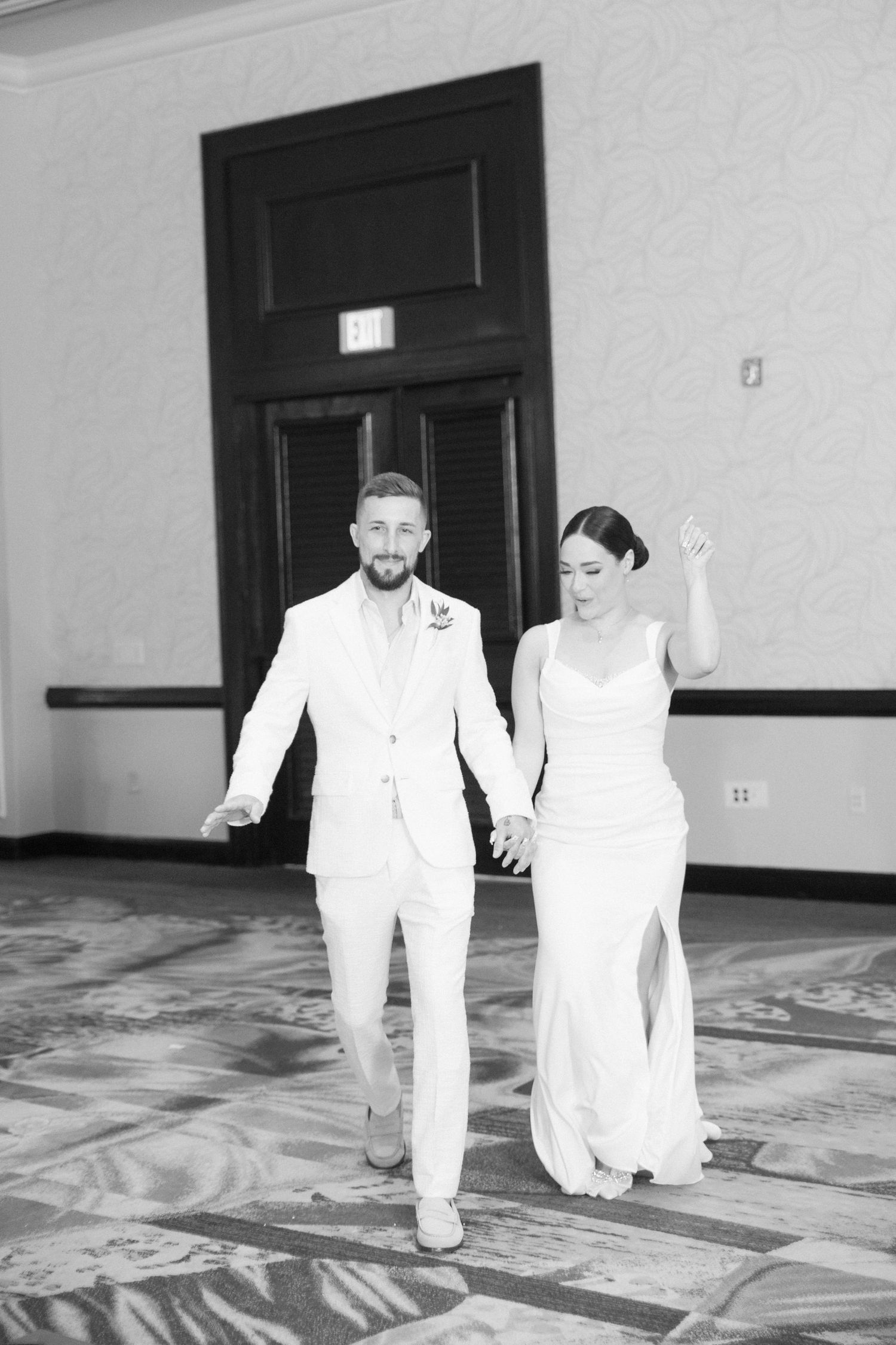 A couple in white attire walks hand in hand through an elegant hallway.