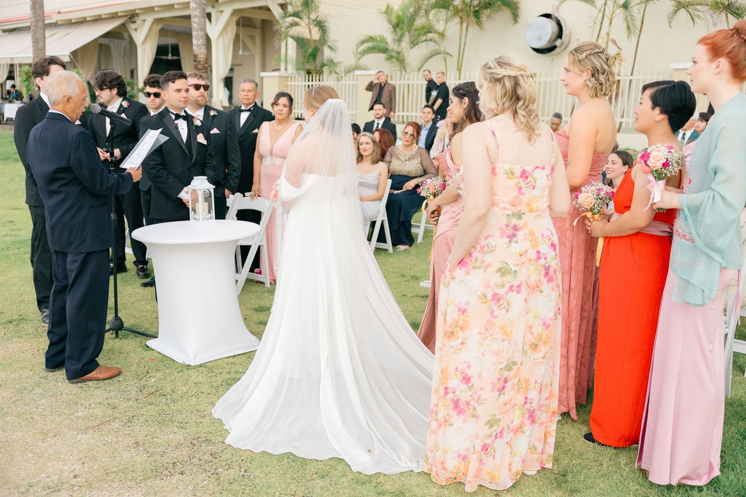 Wedding guests in colorful attire attend outdoor ceremony with palm trees and white chairs.