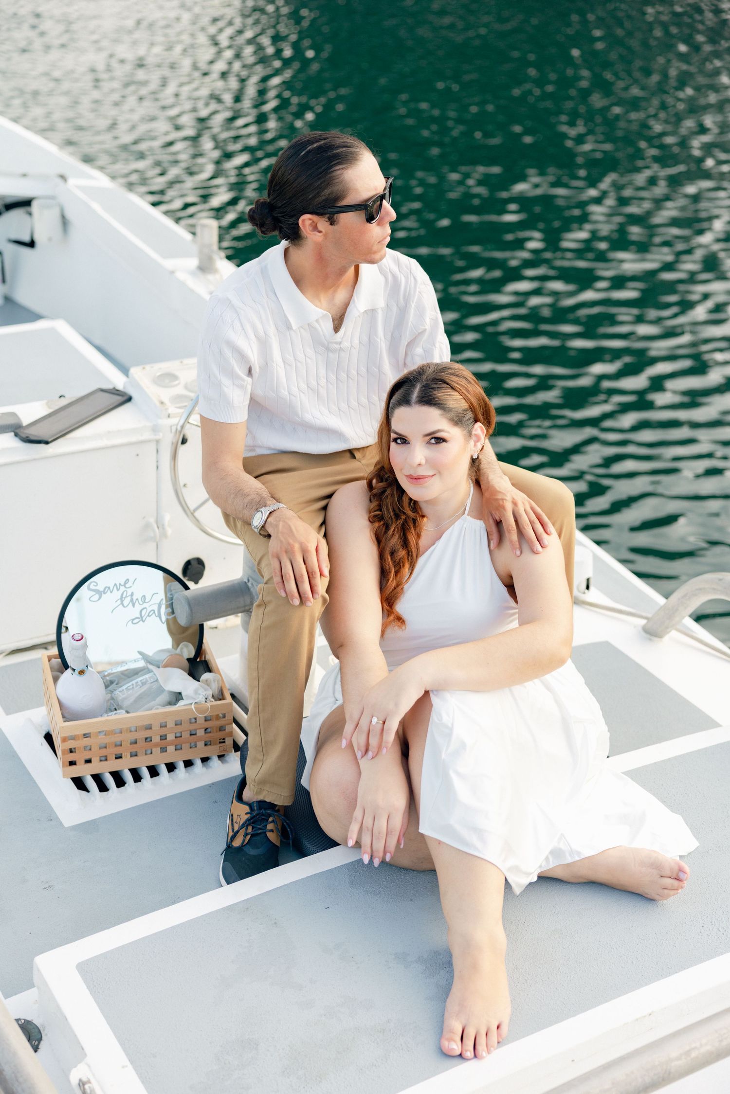 Couple in white and khaki relaxing on yacht deck by emerald waters.