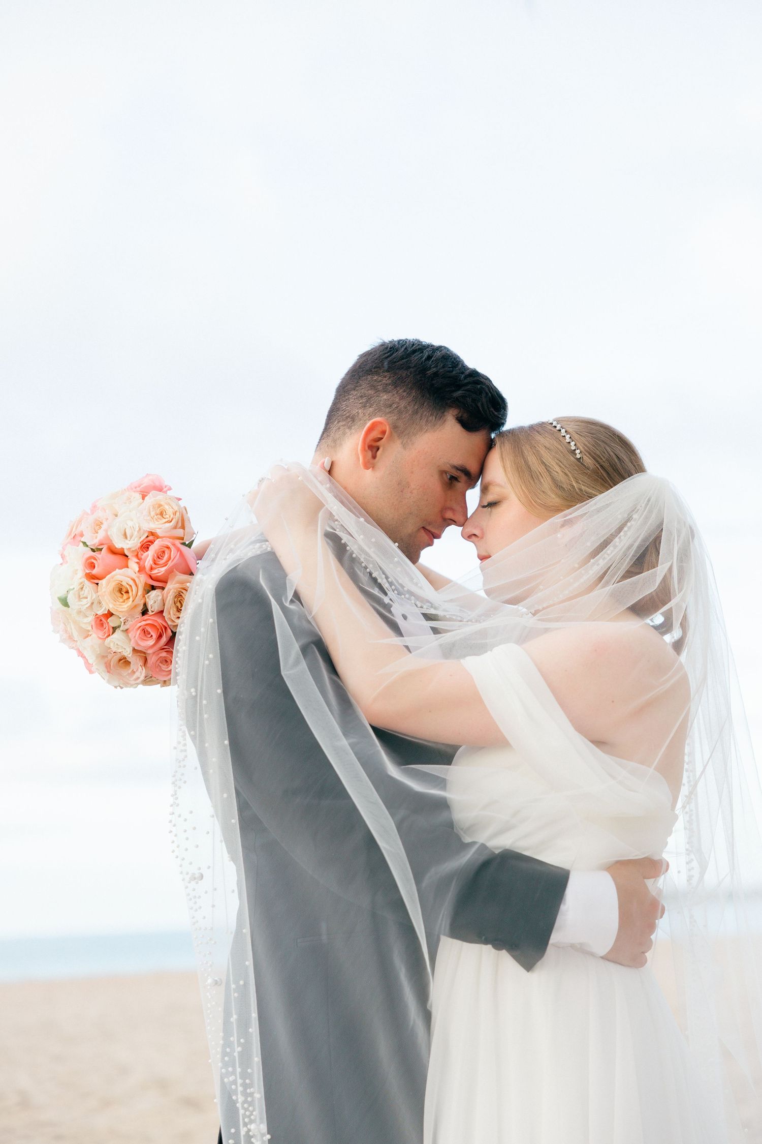Romantic beach wedding couple embracing under a veil with coral bouquet in the background.