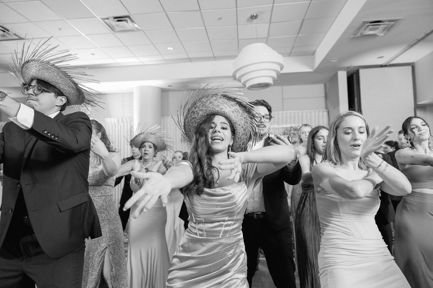 Wedding guests dance energetically in a reception hall with joyful expressions and raised arms.