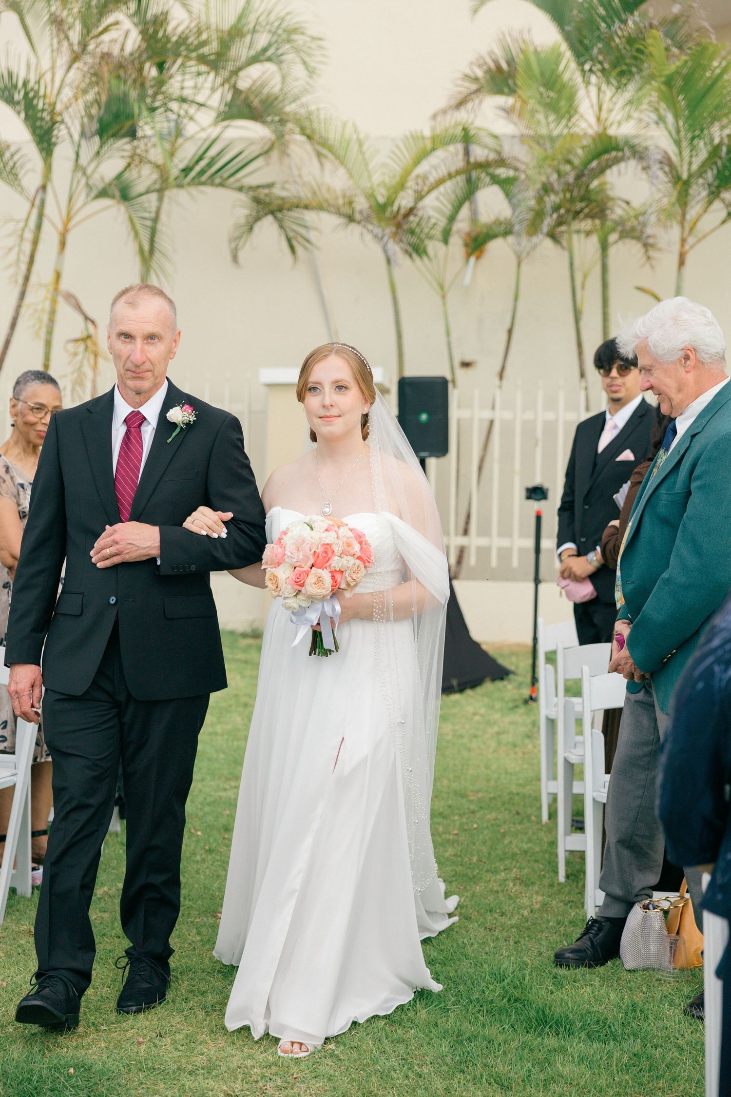 Outdoor wedding ceremony with guests seated on white chairs surrounded by palm trees.
