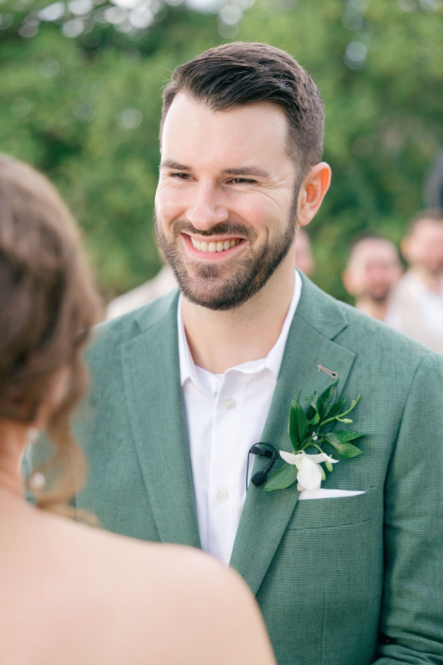 Groom in green suit smiles during outdoor wedding ceremony.