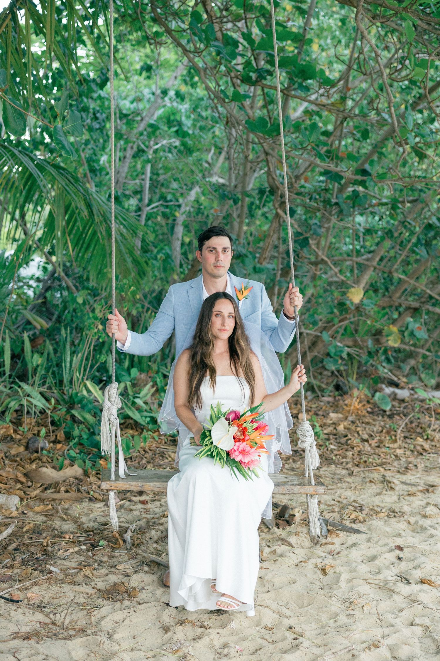 Couple poses on rustic wooden swing surrounded by tropical greenery during wedding photoshoot.