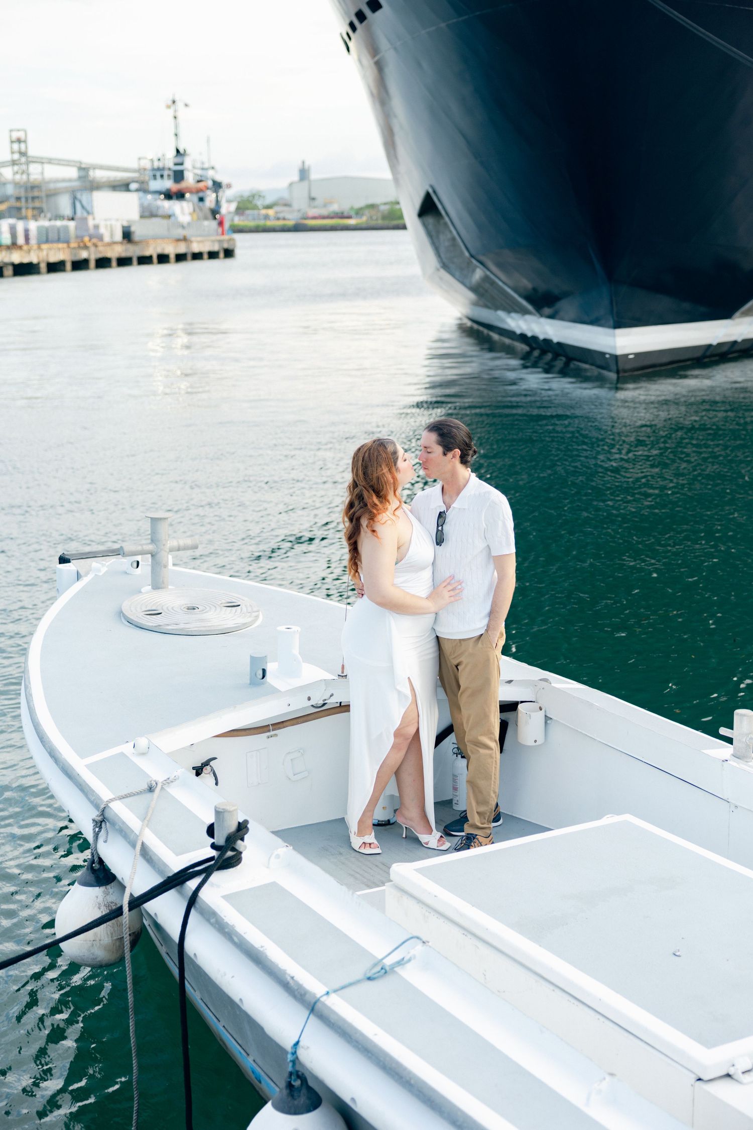 A couple embraces on a pristine white boat with blue water and ships in the background.