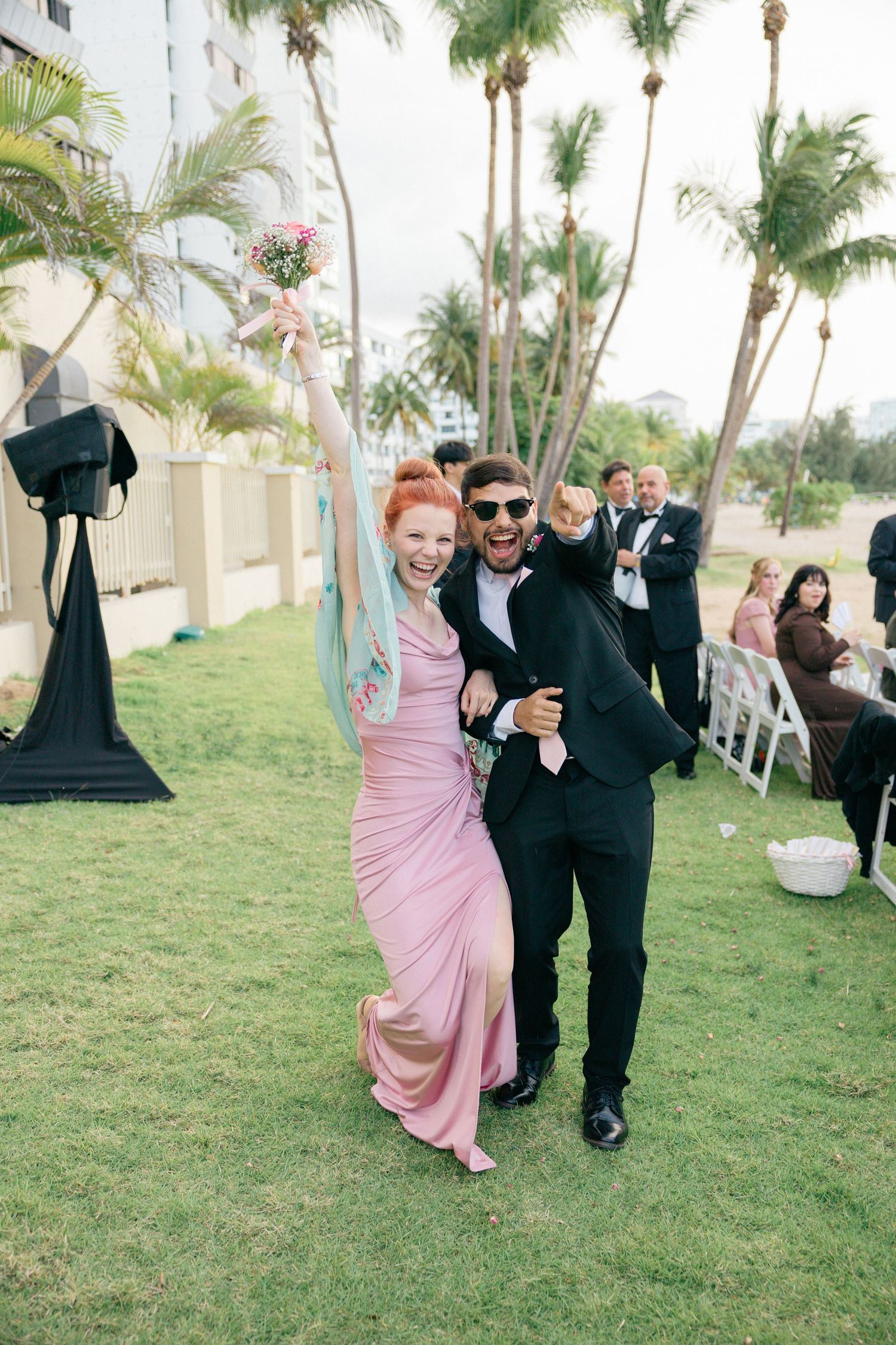 Wedding party members dance energetically on a lawn during an outdoor celebration.
