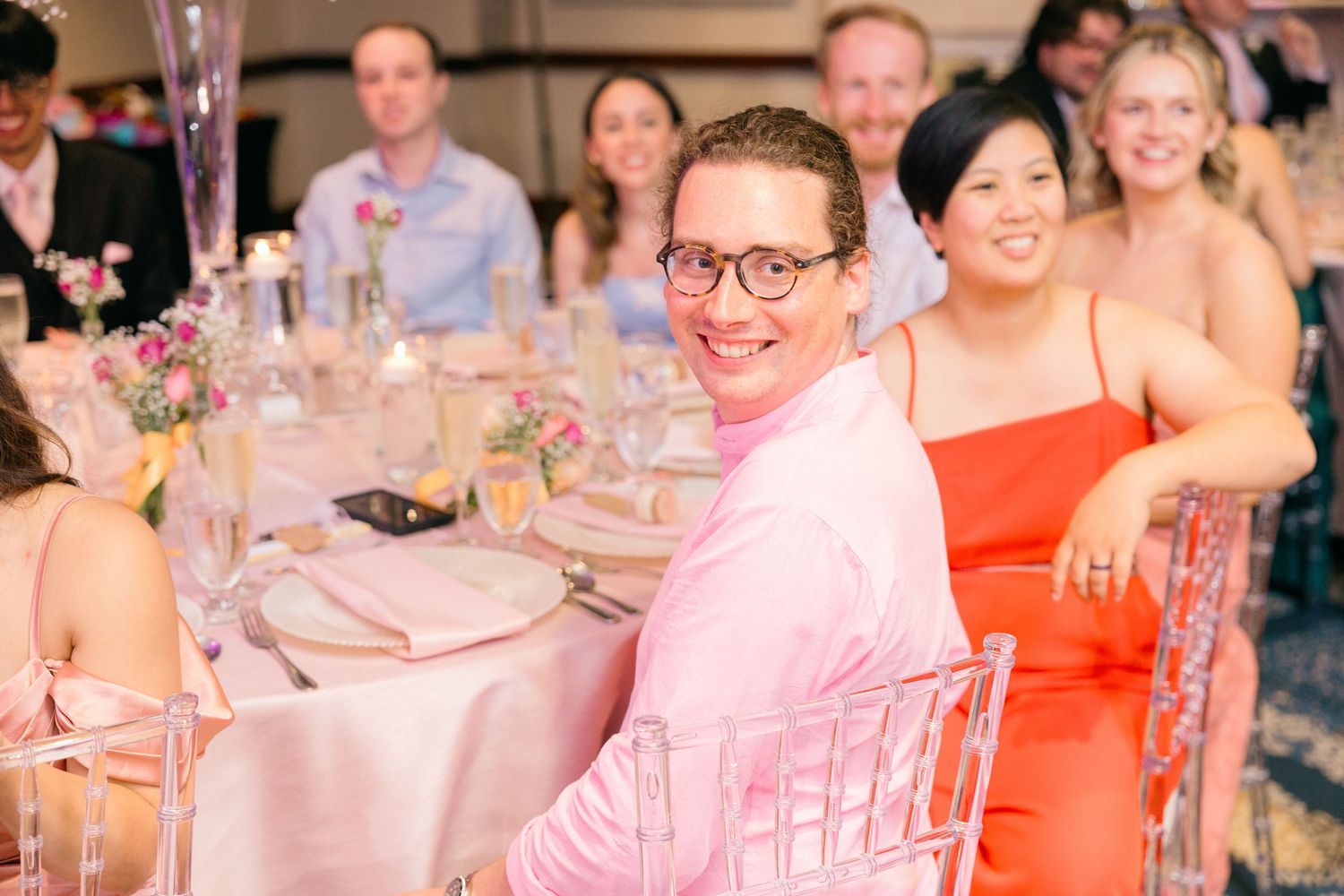 Wedding reception guests seated at round tables with pink decor and floral centerpieces.