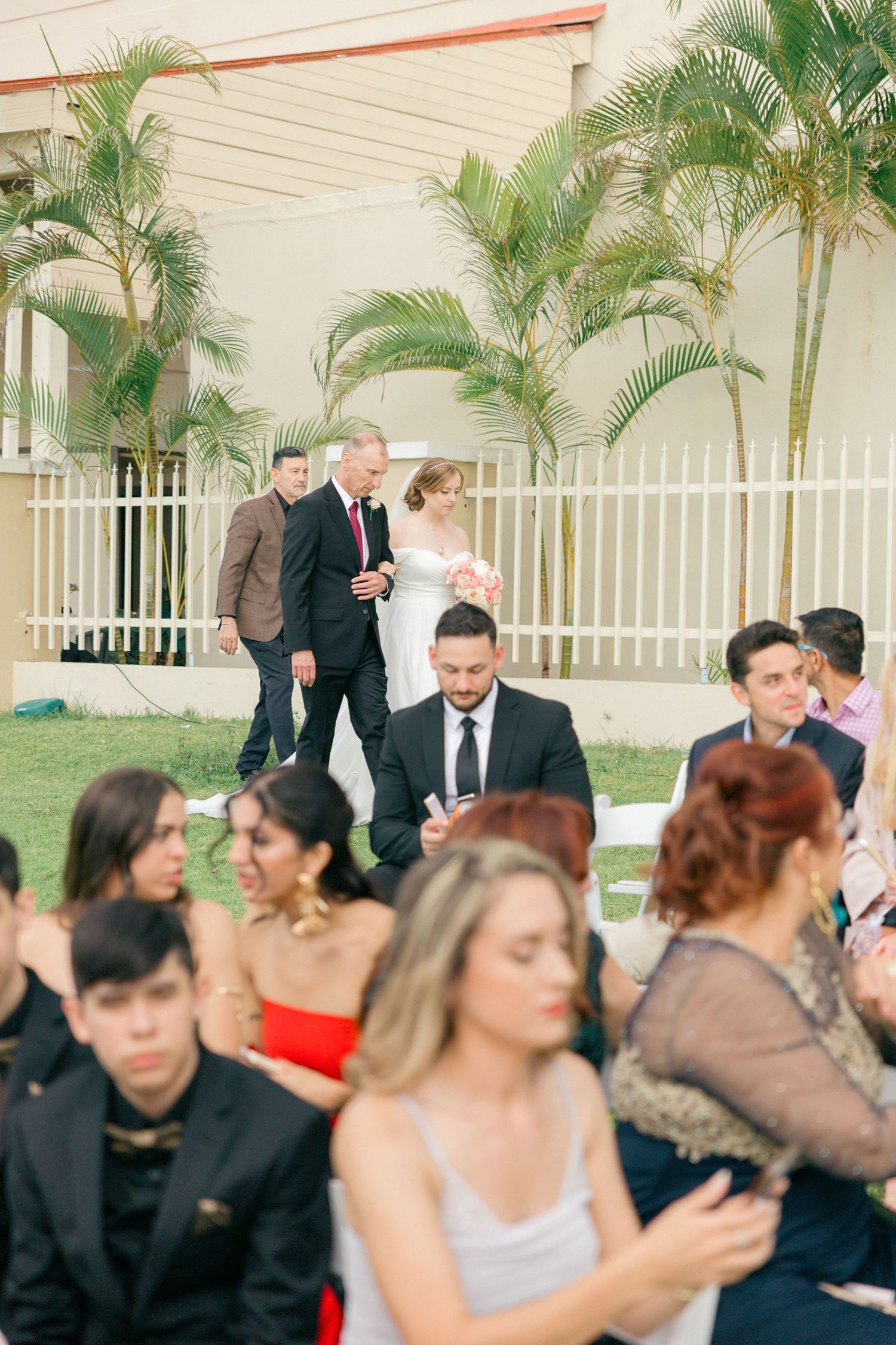 Wedding guests mingle on a lawn with tropical palm trees and beige building exterior.