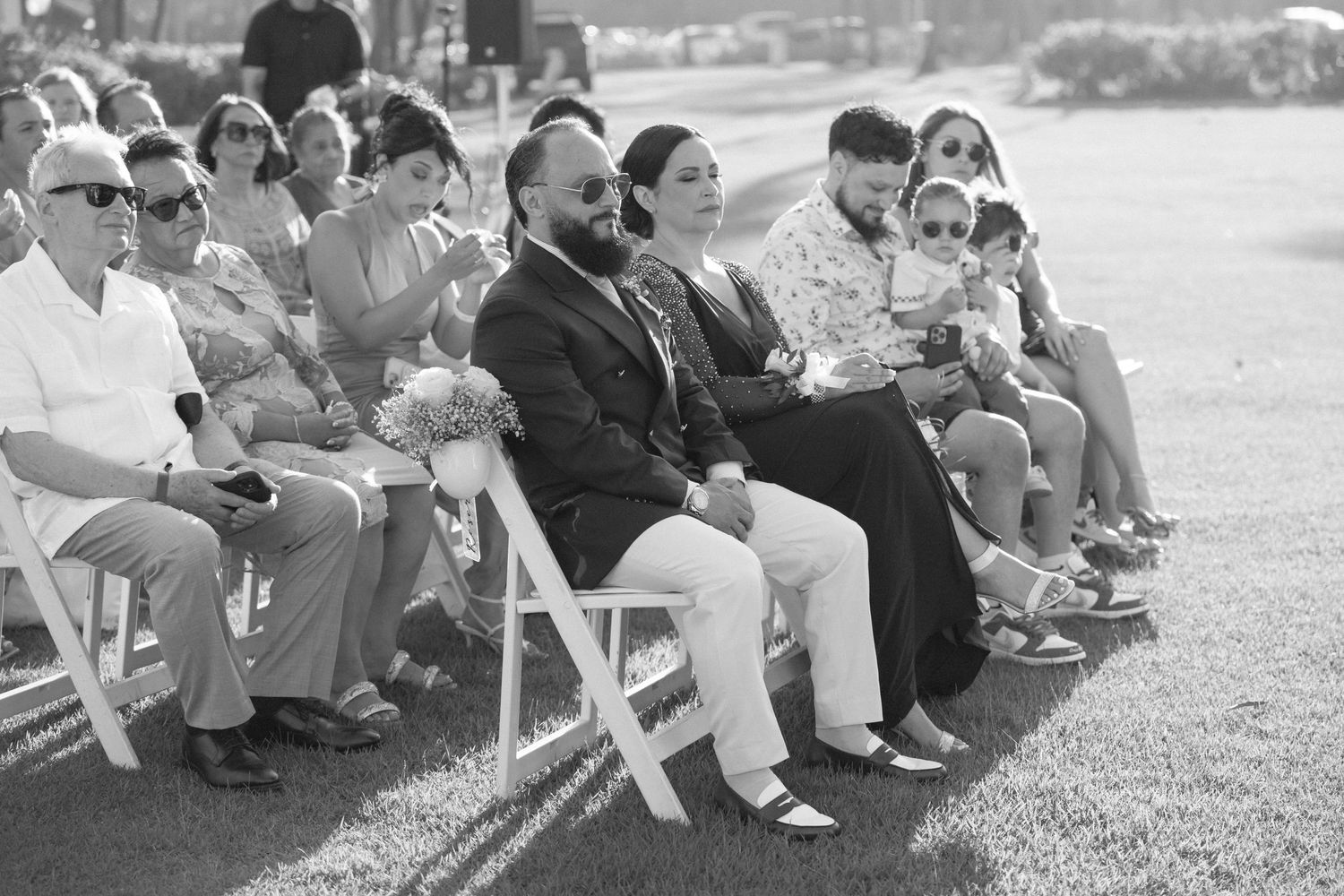 Wedding guests seated on white chairs during an outdoor ceremony.