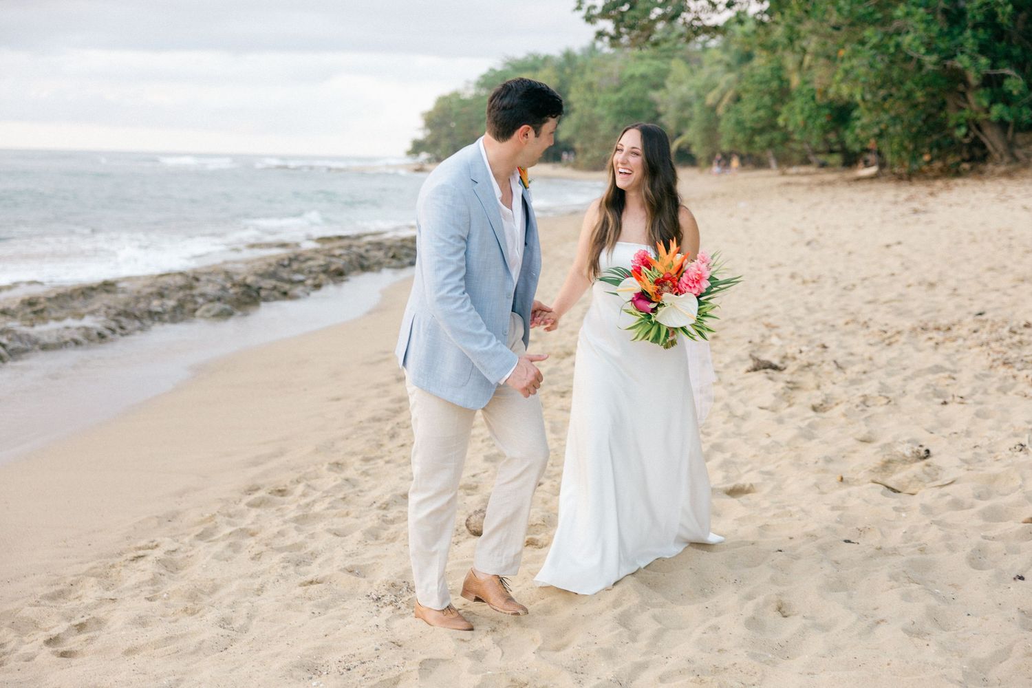 Couple walking hand in hand on a tropical beach wearing light summer wedding attire.