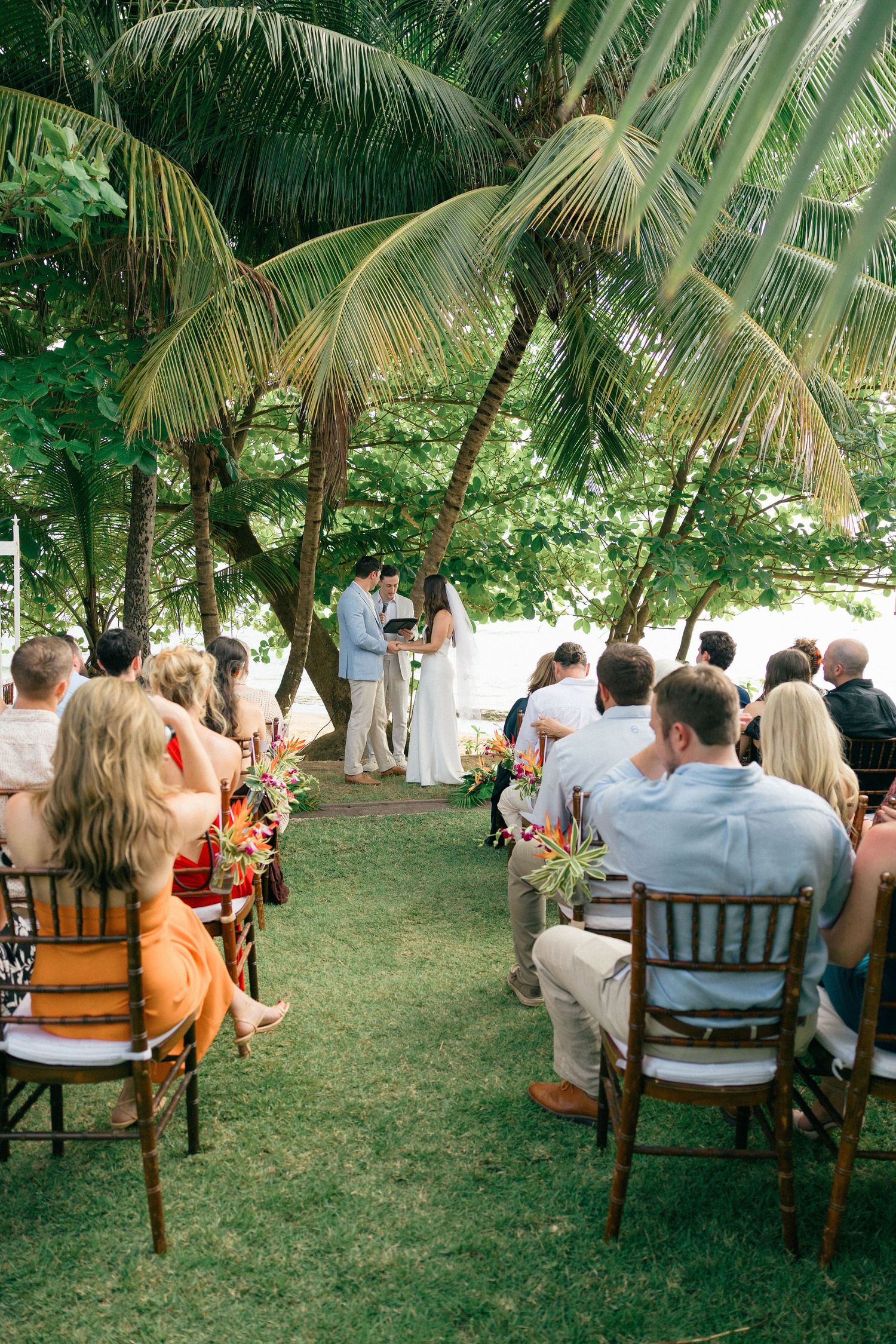 Outdoor wedding ceremony under palm trees with guests seated on wooden chairs on grass lawn.