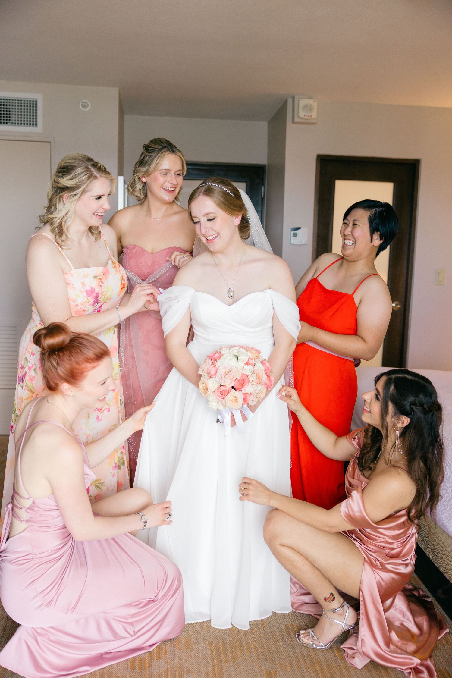 Bridesmaids in pink and coral dresses help with wedding preparations in a hotel room.