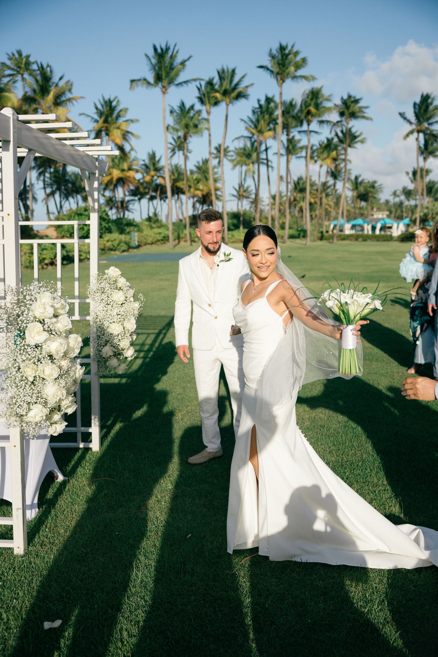 Tropical outdoor wedding ceremony with white decor and palm trees against a bright blue sky.