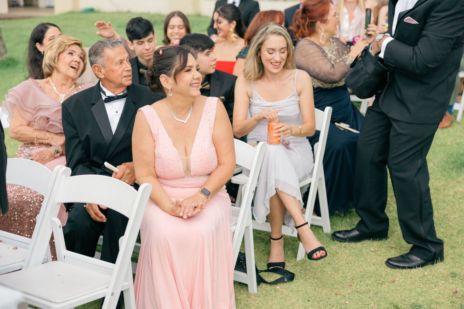 Wedding guests in formal attire gather outdoors on a grassy lawn with palm trees and a beige building in the background.