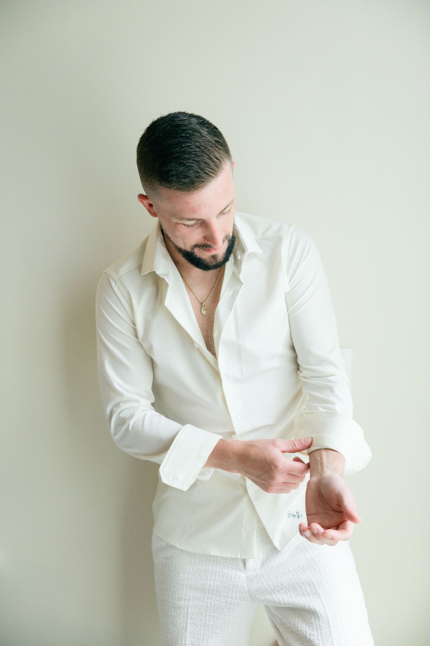Stylish pose in white button-down shirt and pants against light wall.