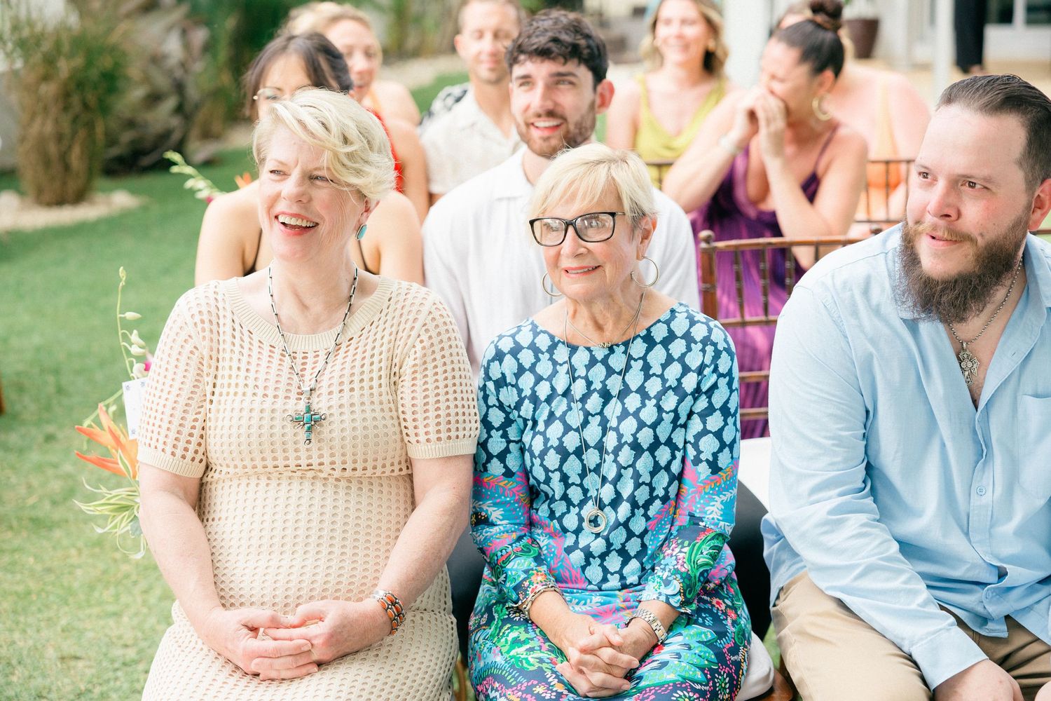 Wedding guests in casual attire smile while seated outdoors during tropical ceremony.