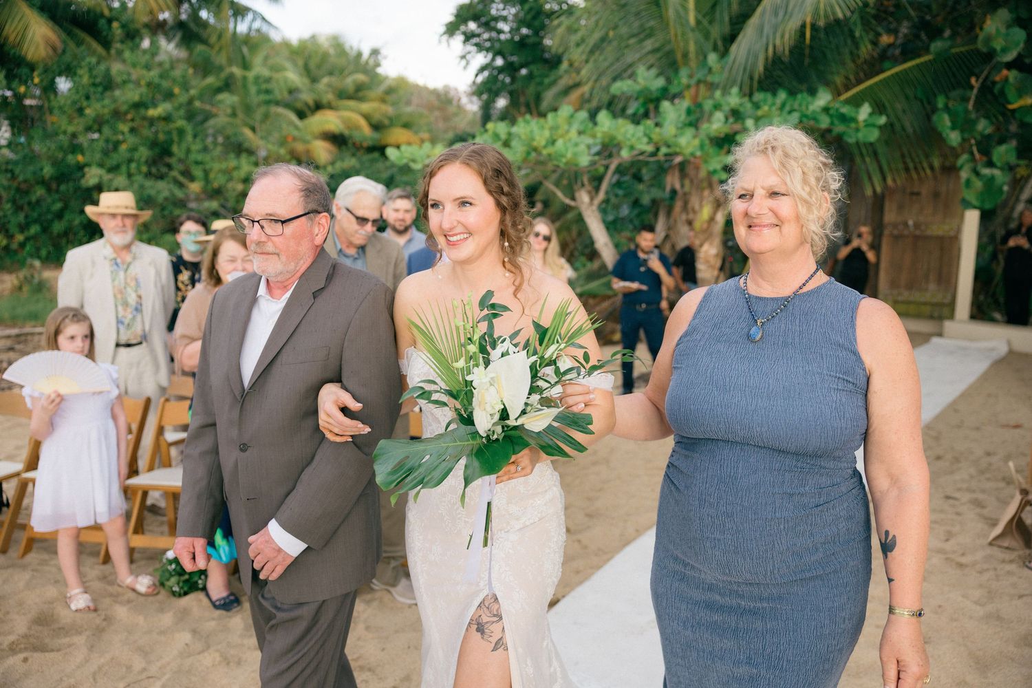 Wedding ceremony participants gather on sandy beach with tropical foliage backdrop.