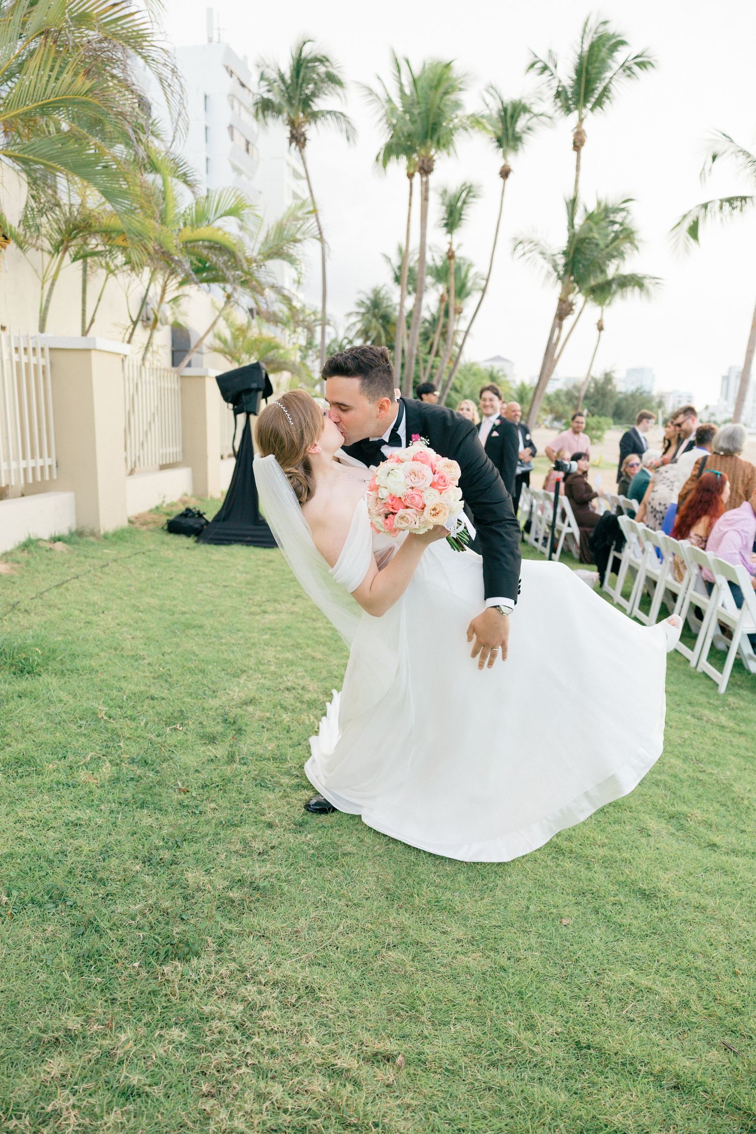 Wedding celebration on a grassy lawn with palm trees and guests in formal attire.