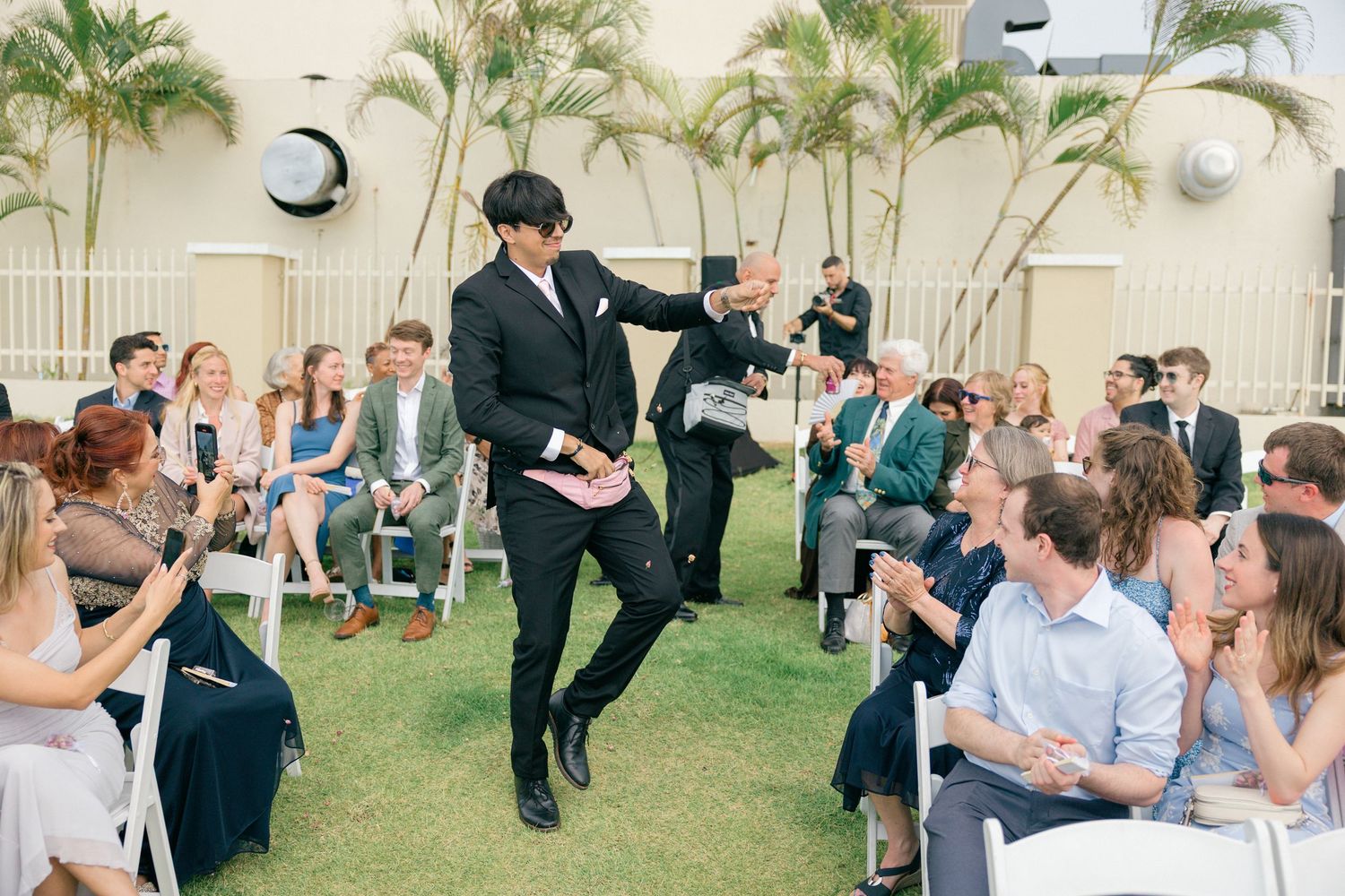 Outdoor wedding venue setup with white chairs and tropical palm trees against a light colored wall.
