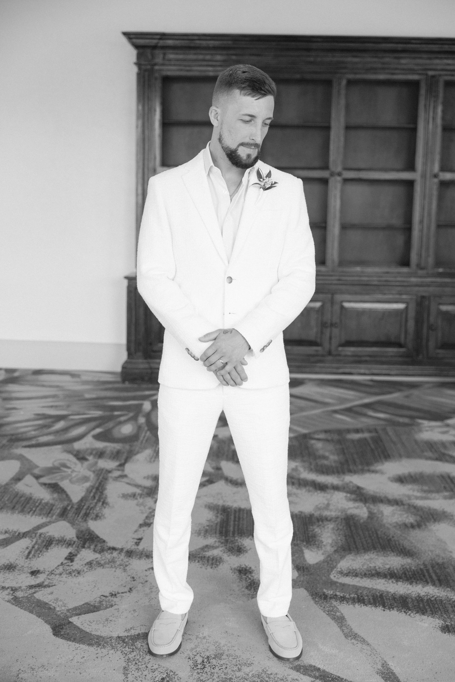 Groom in white suit poses on patterned floor against wooden paneled wall.