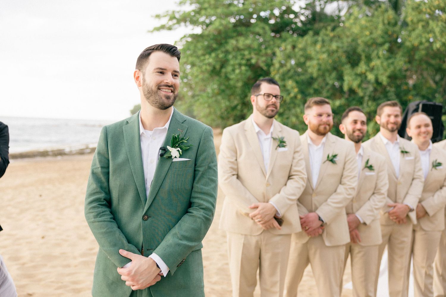 Wedding party members in beige suits and green jacket stand together on beach.