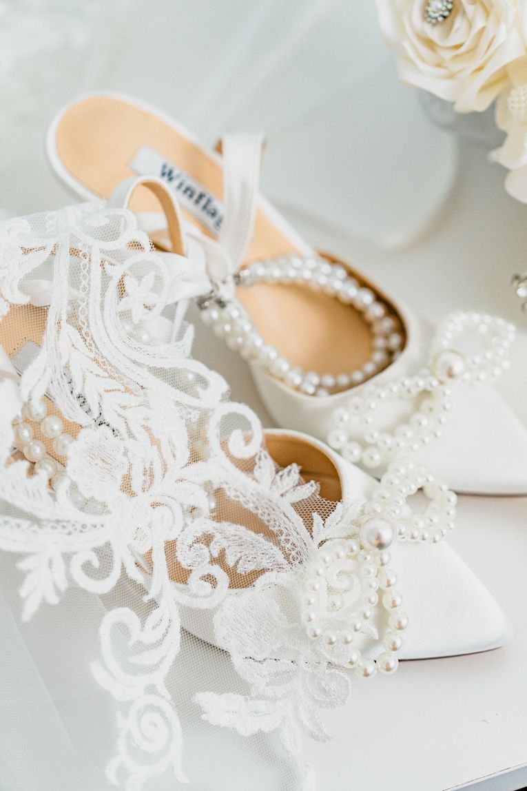 Ornate white lace bridal shoes with pearl details on light background.