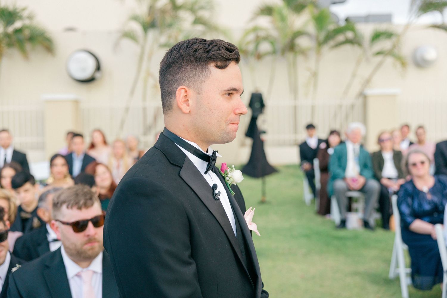 Palm trees frame an outdoor wedding ceremony with guests in formal attire.