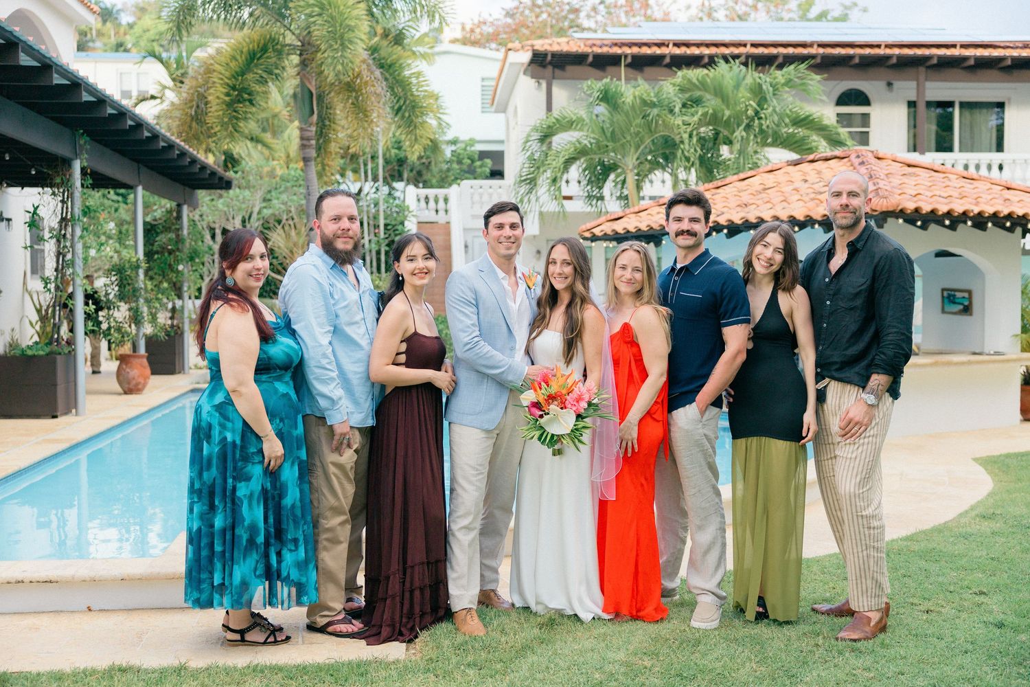 Wedding party poses together in front of a tropical pool area wearing coordinated dresses in shades of blue and coral.