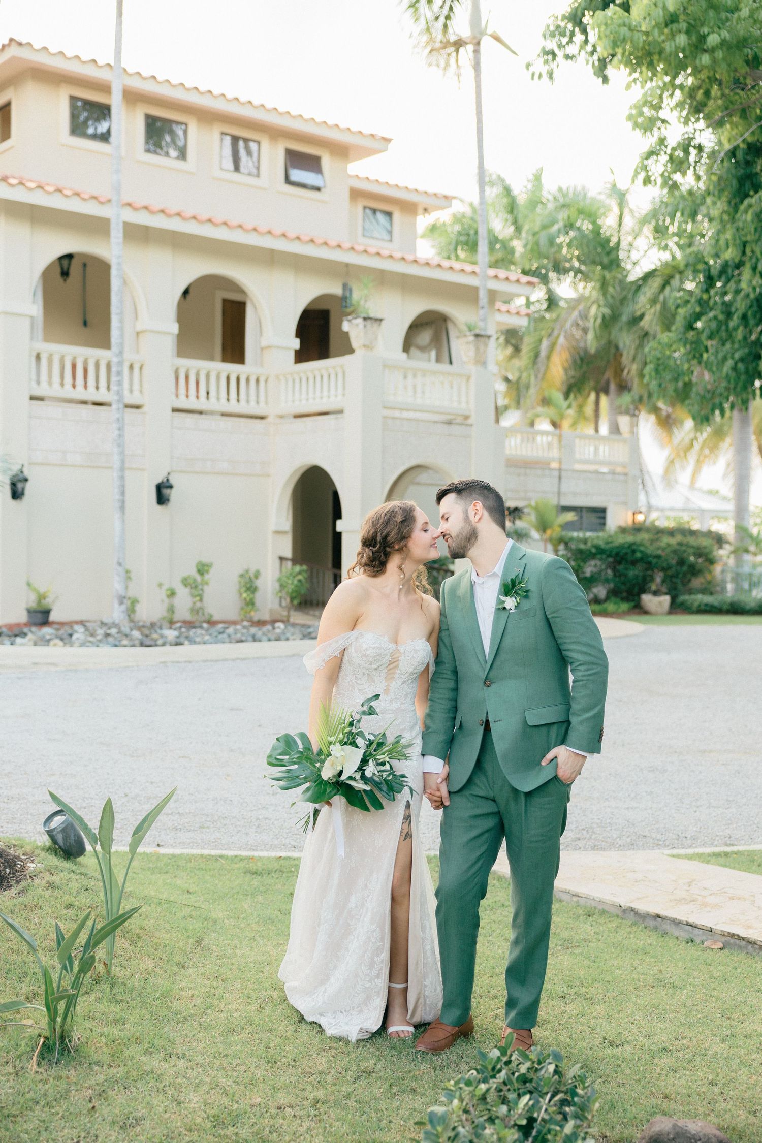 Wedding couple poses in front of Mediterranean-style building with palm trees and manicured lawn.