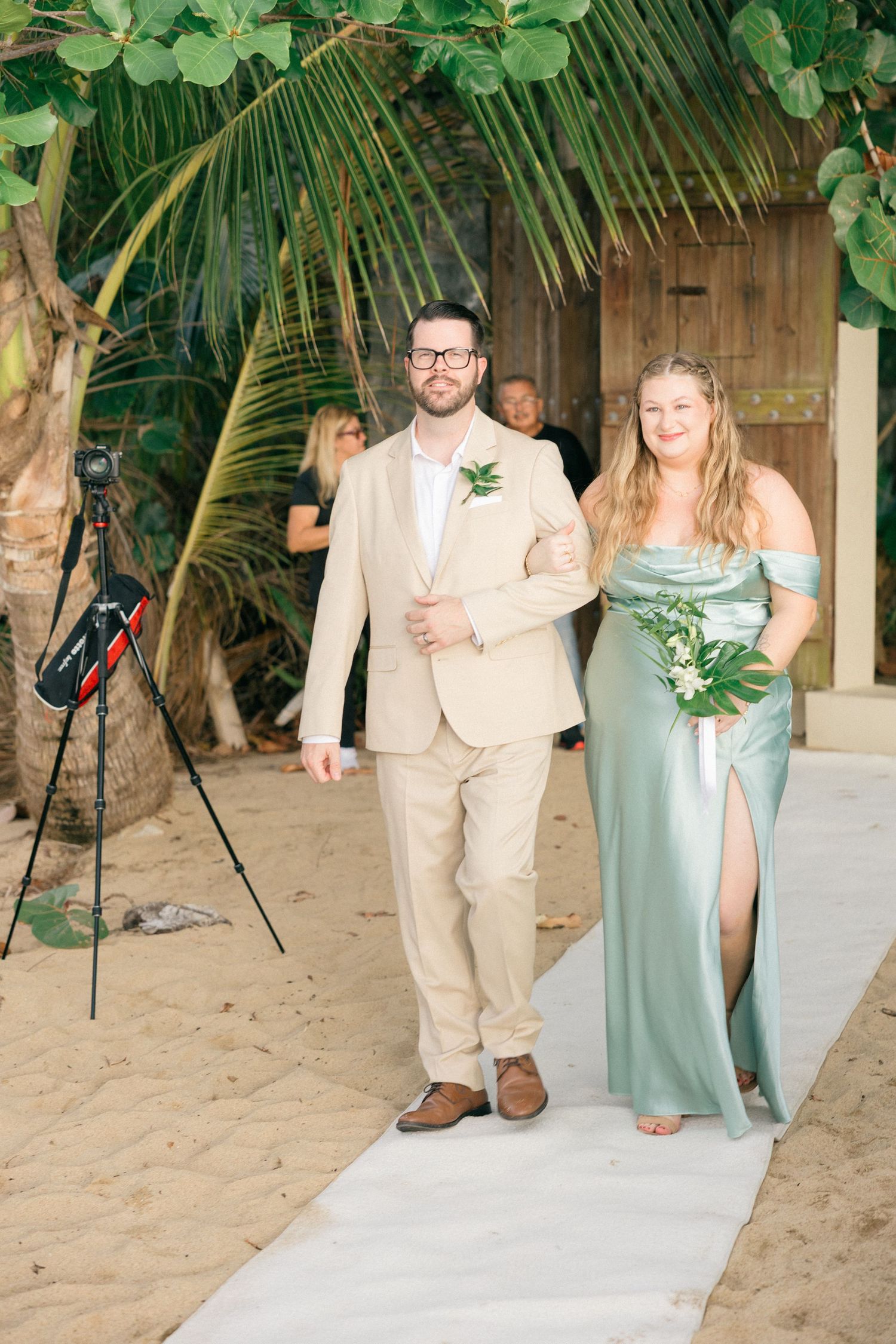 Wedding party members in sage green and beige attire walking down white aisle runner on beach.