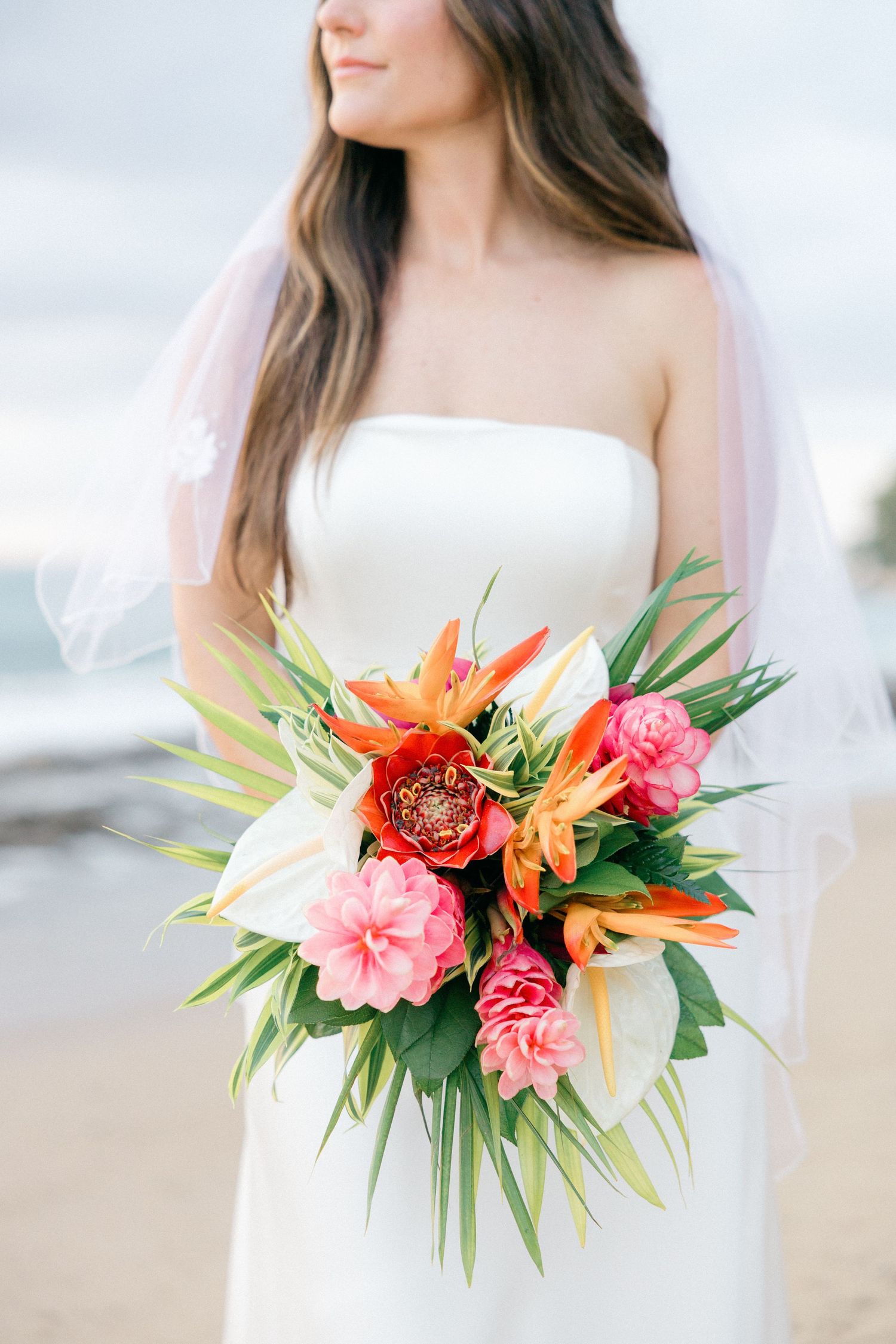 Tropical bridal bouquet featuring orange birds of paradise and pink flowers against a white dress.