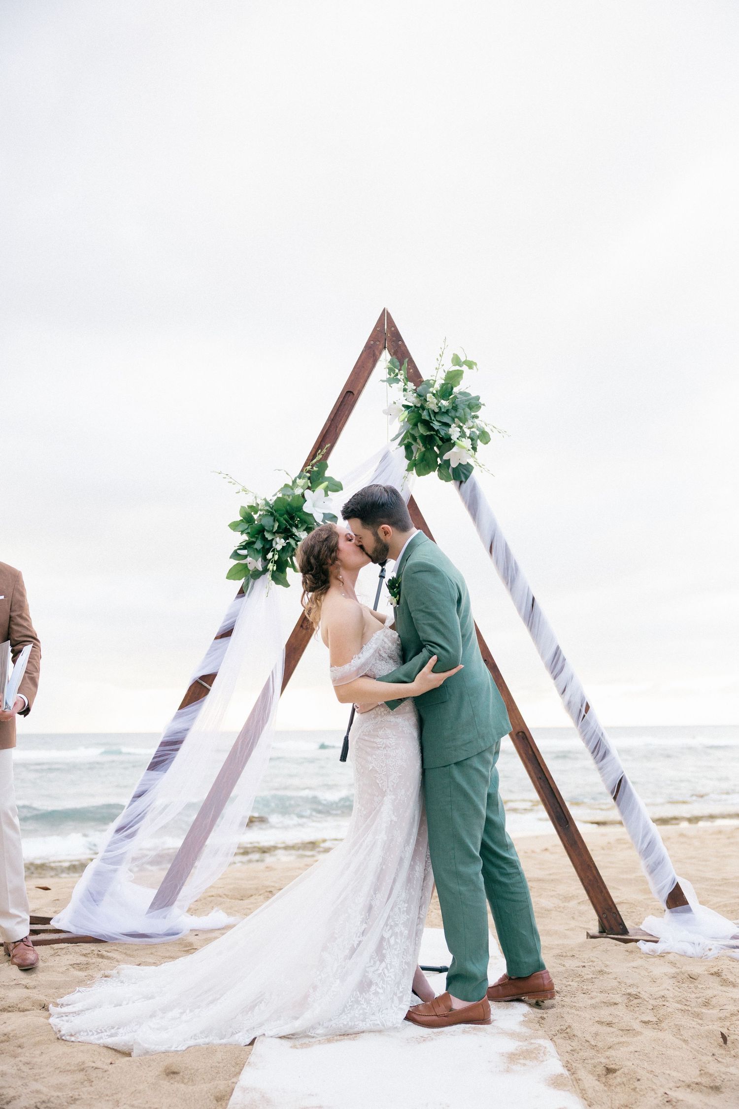 Romantic beach wedding moment under wooden arch with flowing white fabric and greenery.