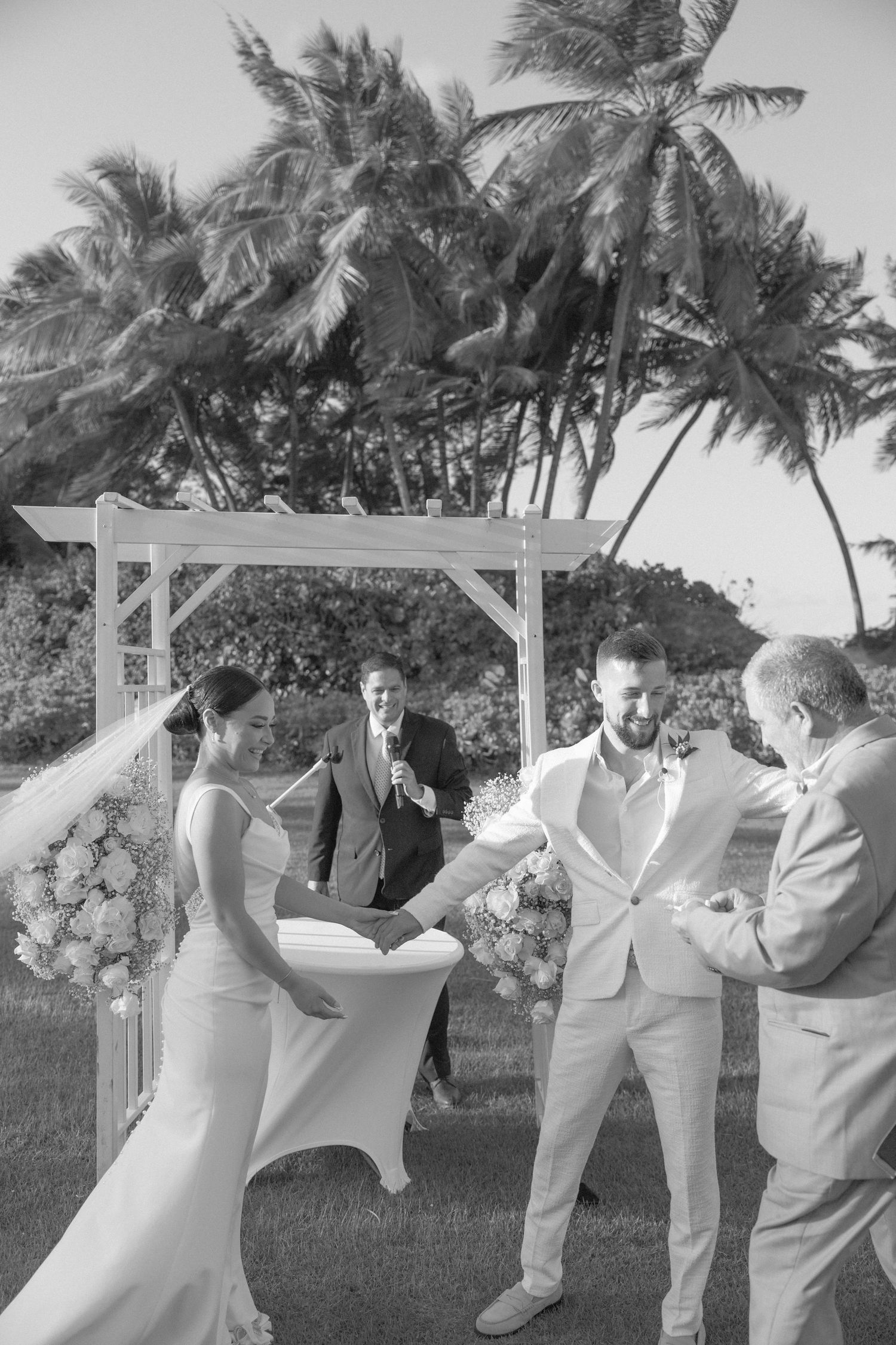 A romantic wedding ceremony moment under a white arbor decorated with flowers and palm trees in the background.