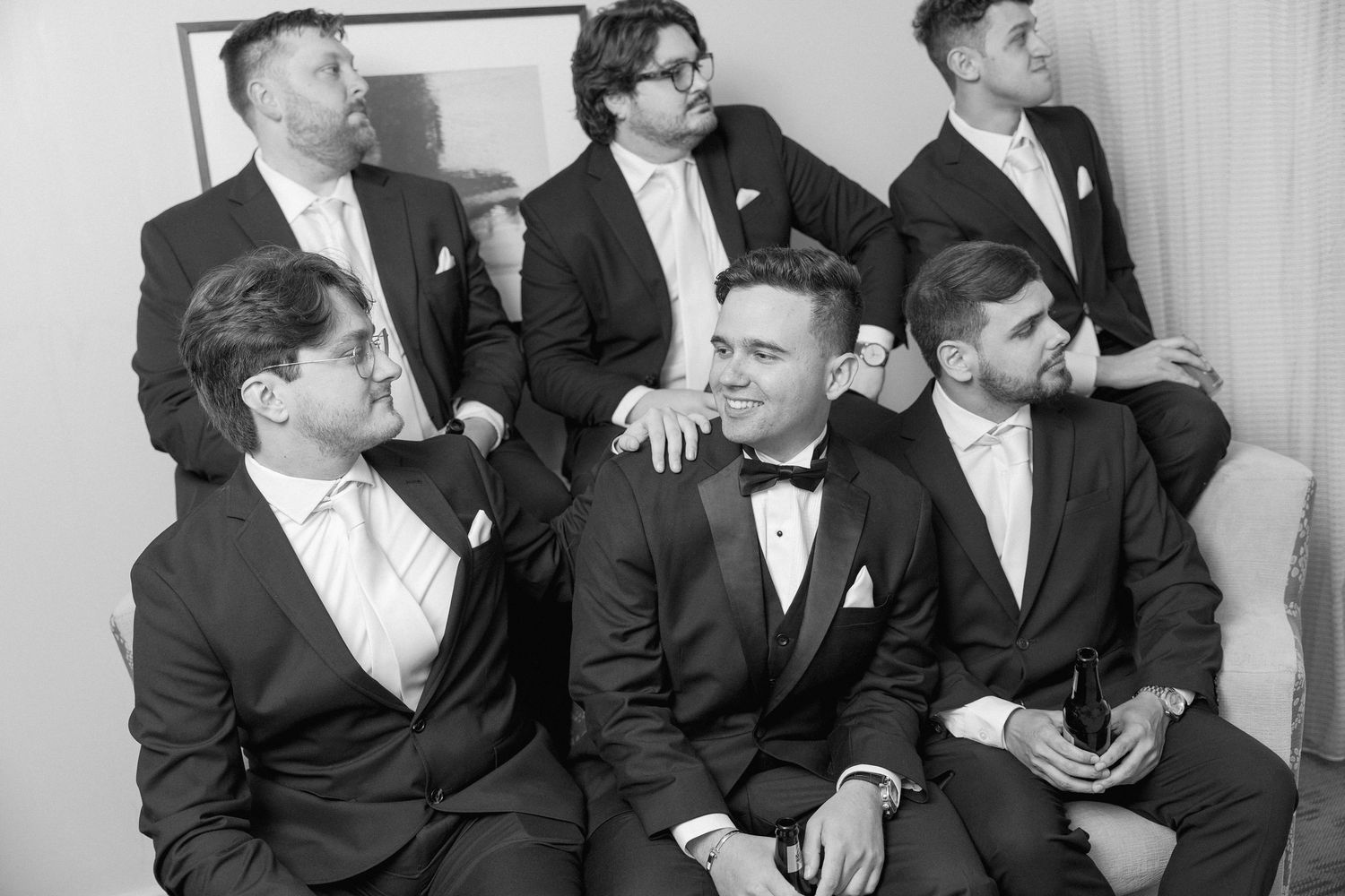 Groomsmen in formal black tuxedos share laughs while posing for group photos at a wedding.